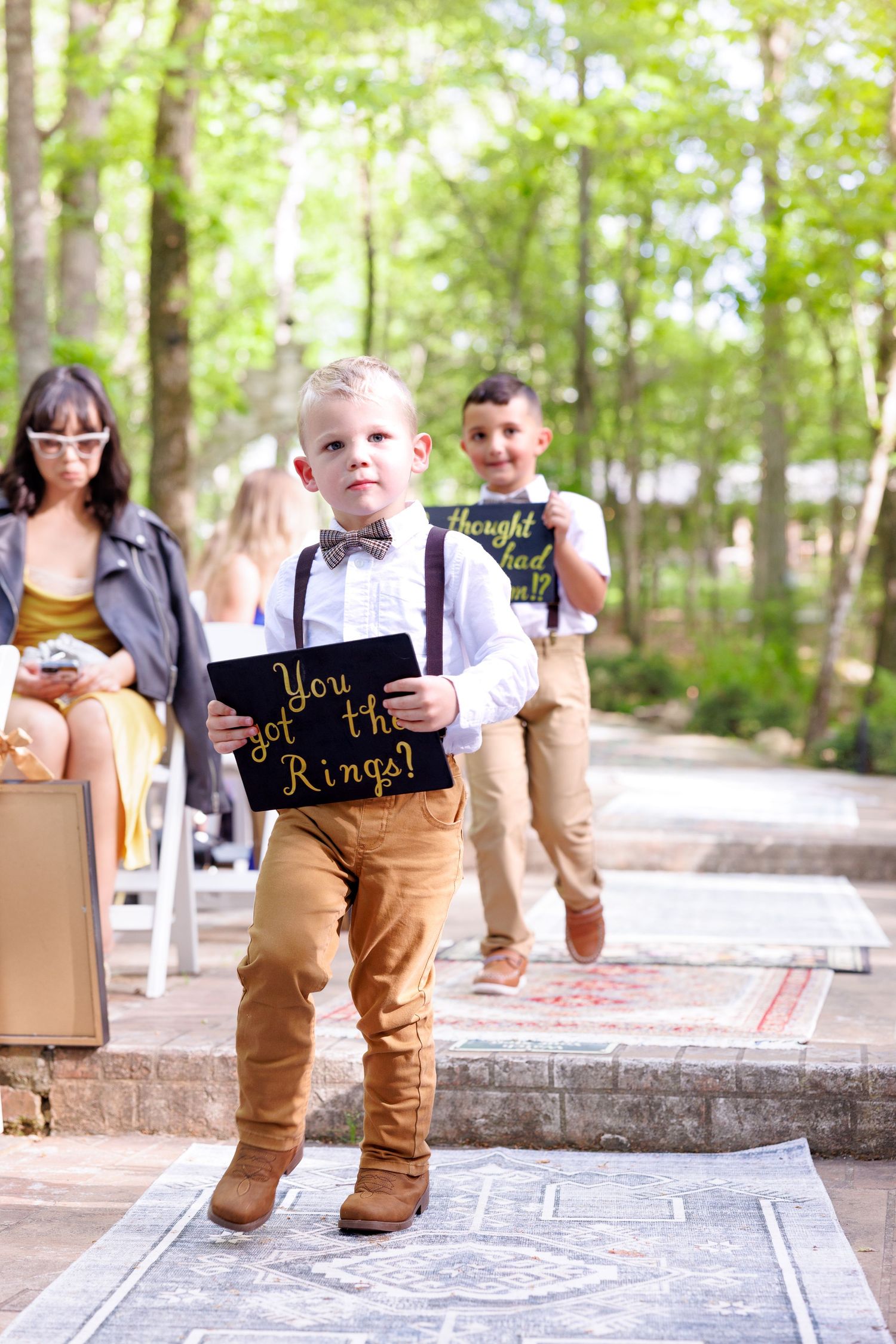 Young ring bearer walks down wedding aisle holding a chalkboard sign reading You Got the Rings at outdoor ceremony.
