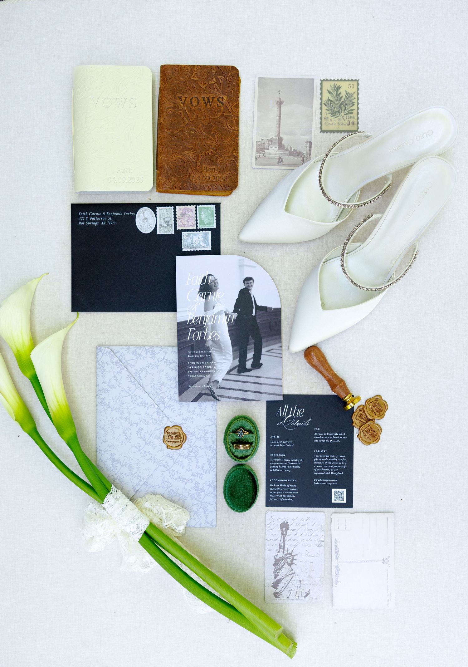 Elegant wedding flat lay with invitation suite, white heels, wax seals, vow books, stamps, and calla lilies on white background.
