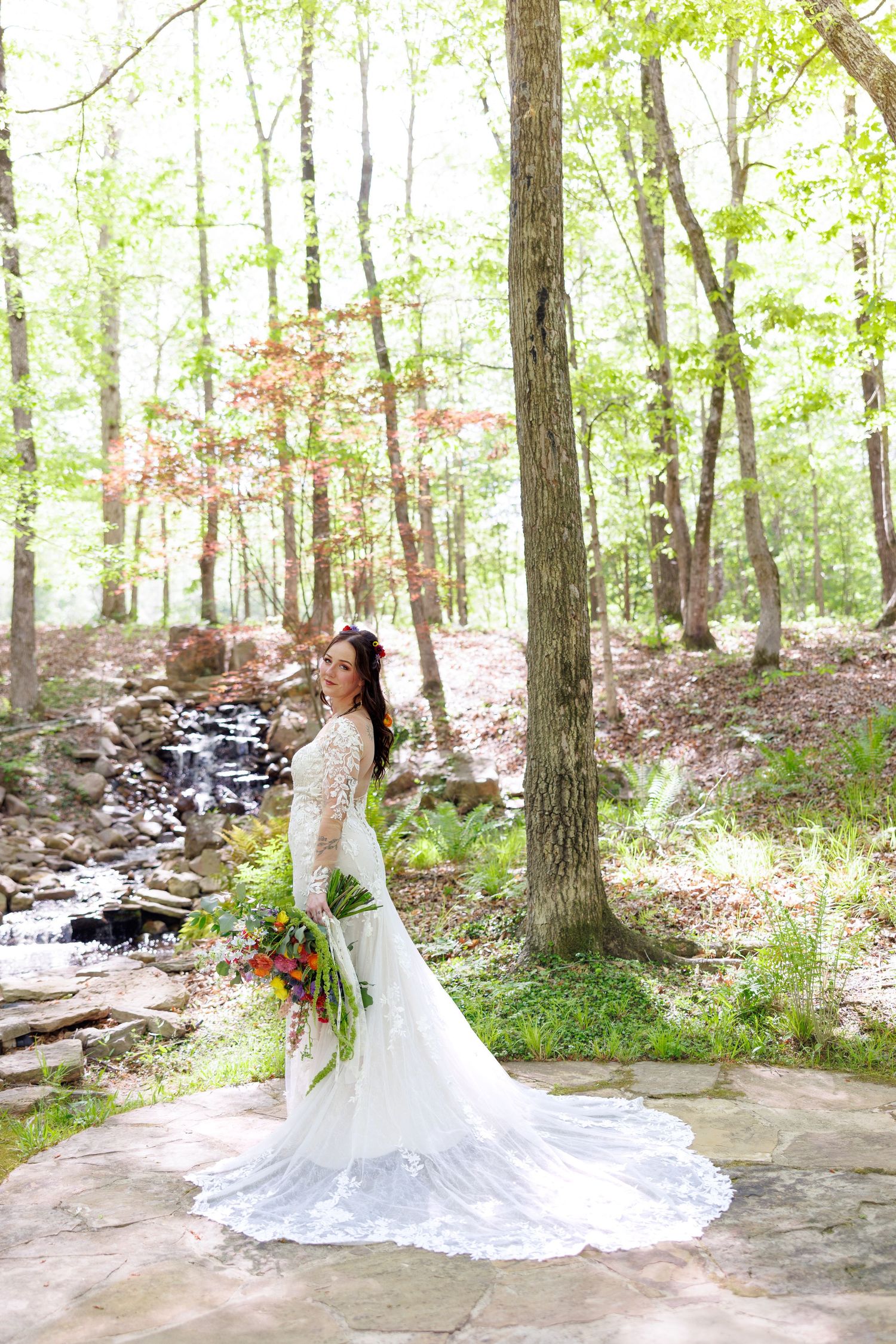 Elegant bride in lace mermaid gown with long train stands in sunlit forest near a stone waterfall.