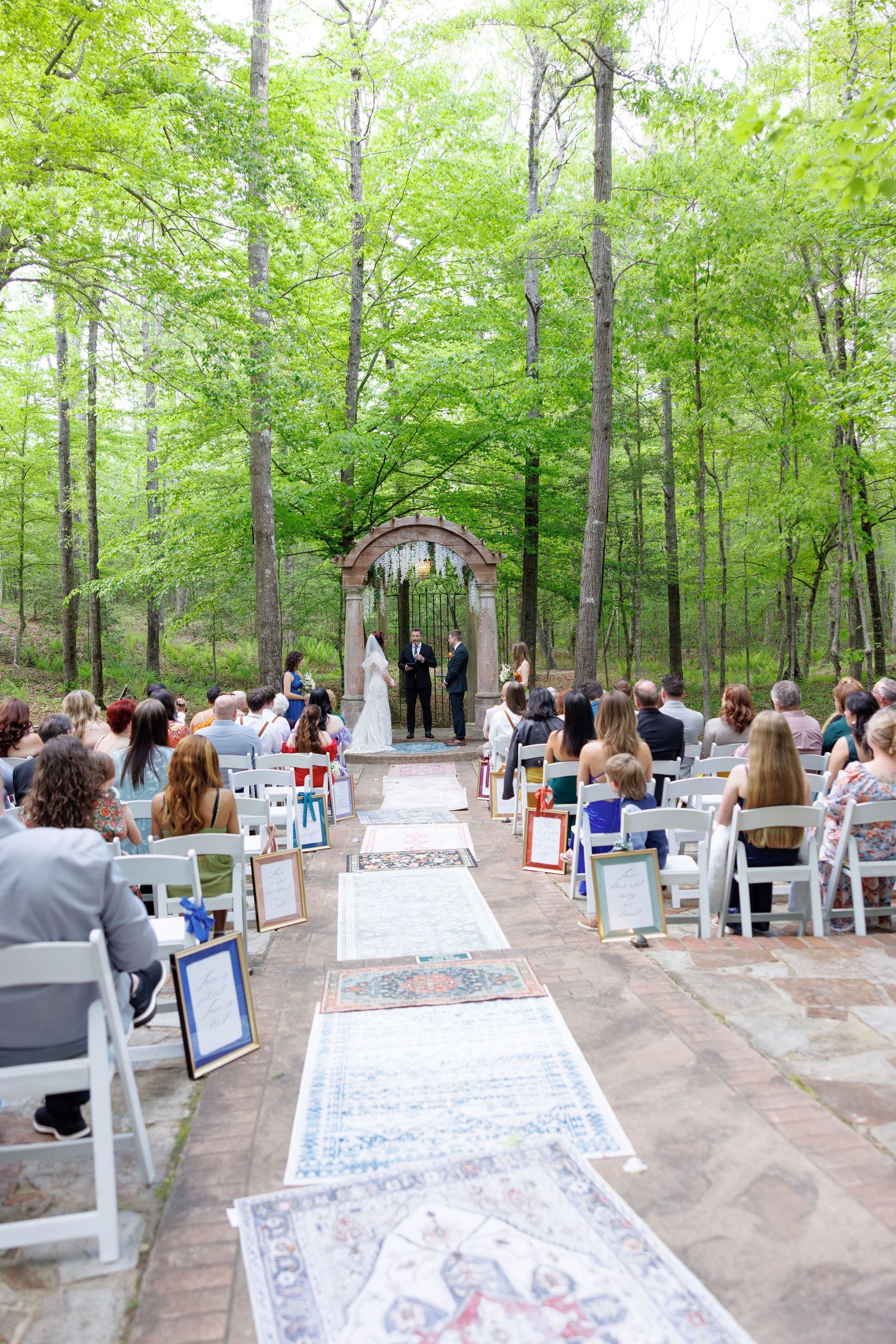 Outdoor wedding ceremony in a lush forest setting with white chairs lining a brick aisle leading to an elegant arch.