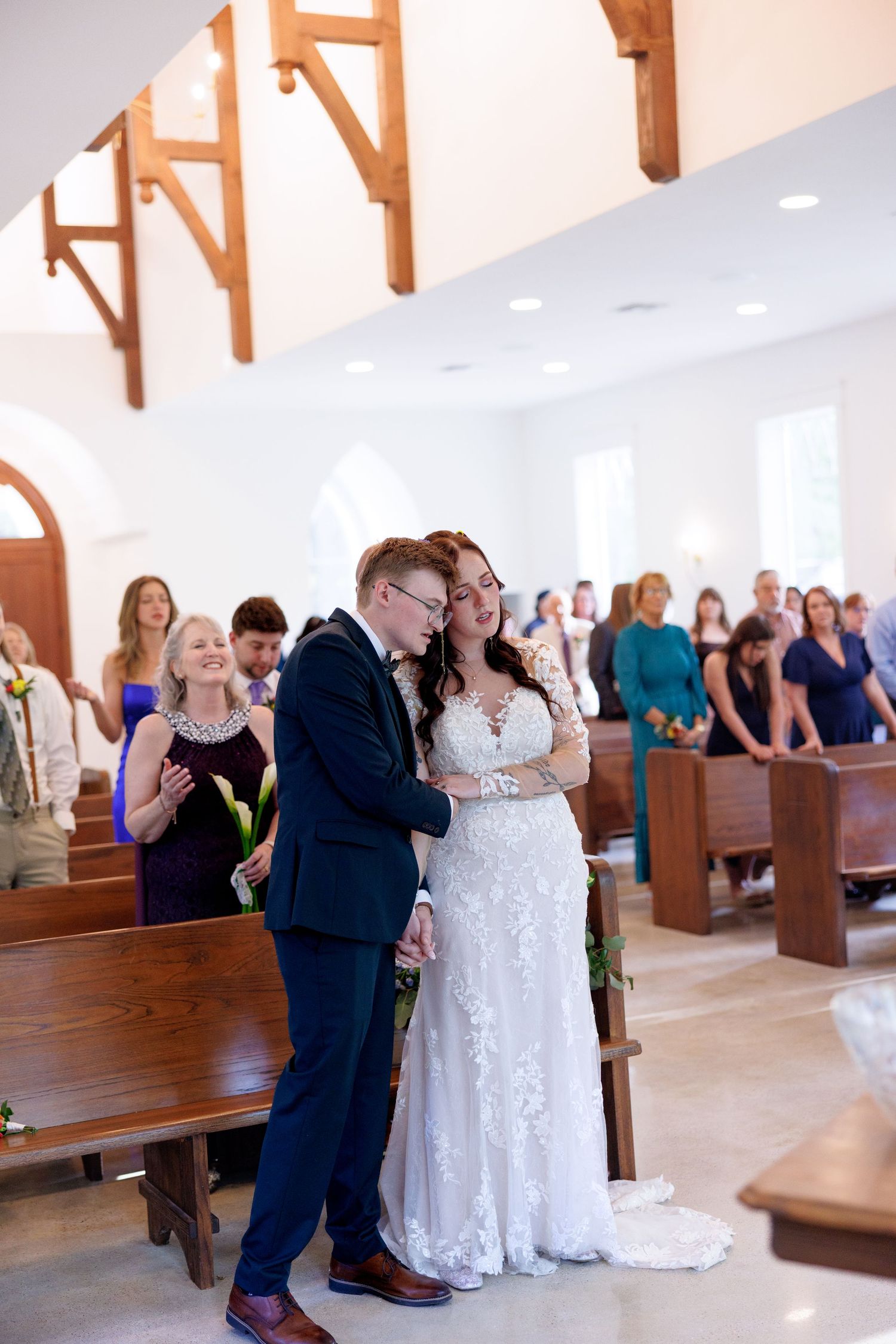 Bride and groom share tender embrace walking down church aisle after wedding ceremony with guests applauding.