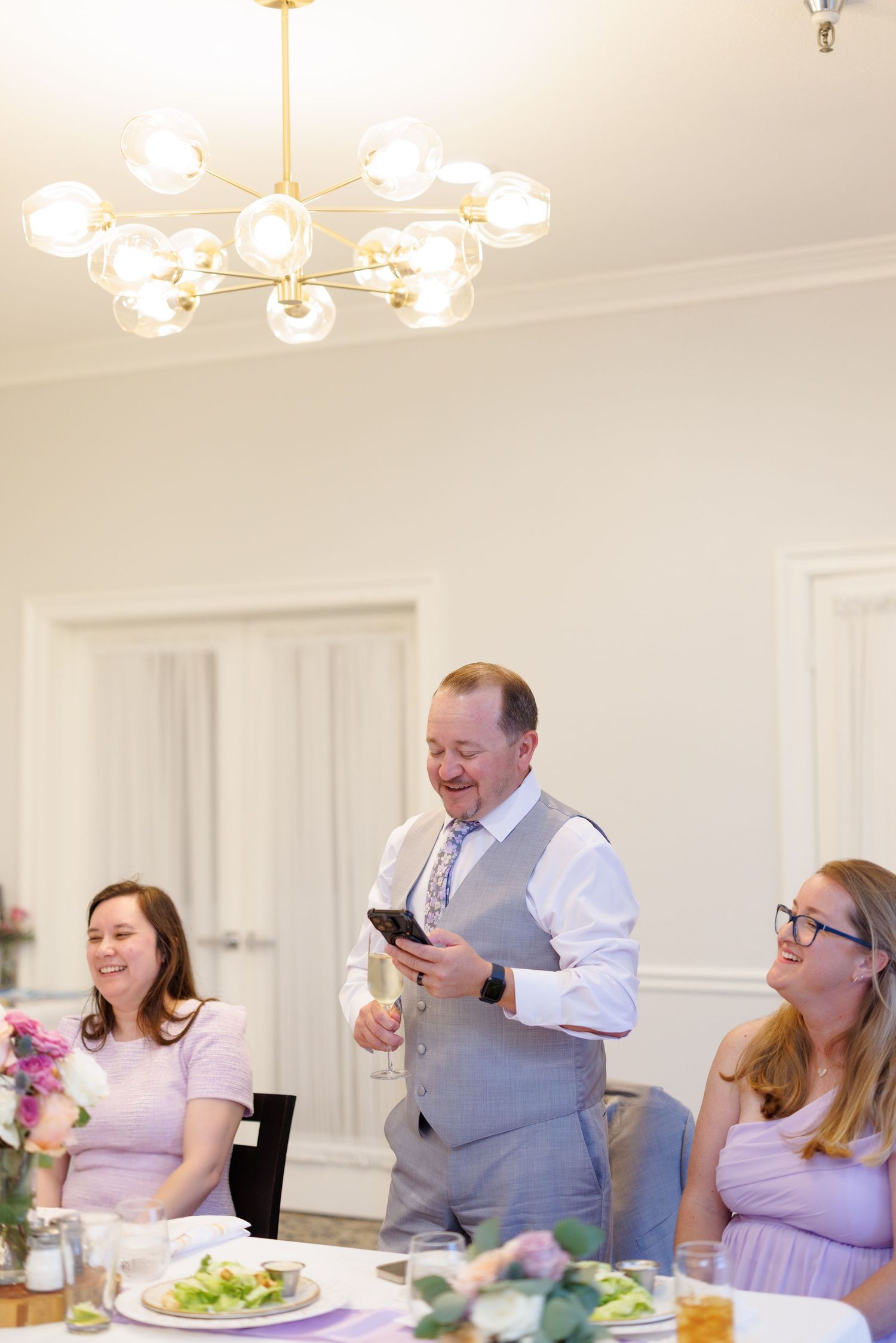 Man in gray vest gives speech holding phone at wedding reception while guests laugh at decorated table.
