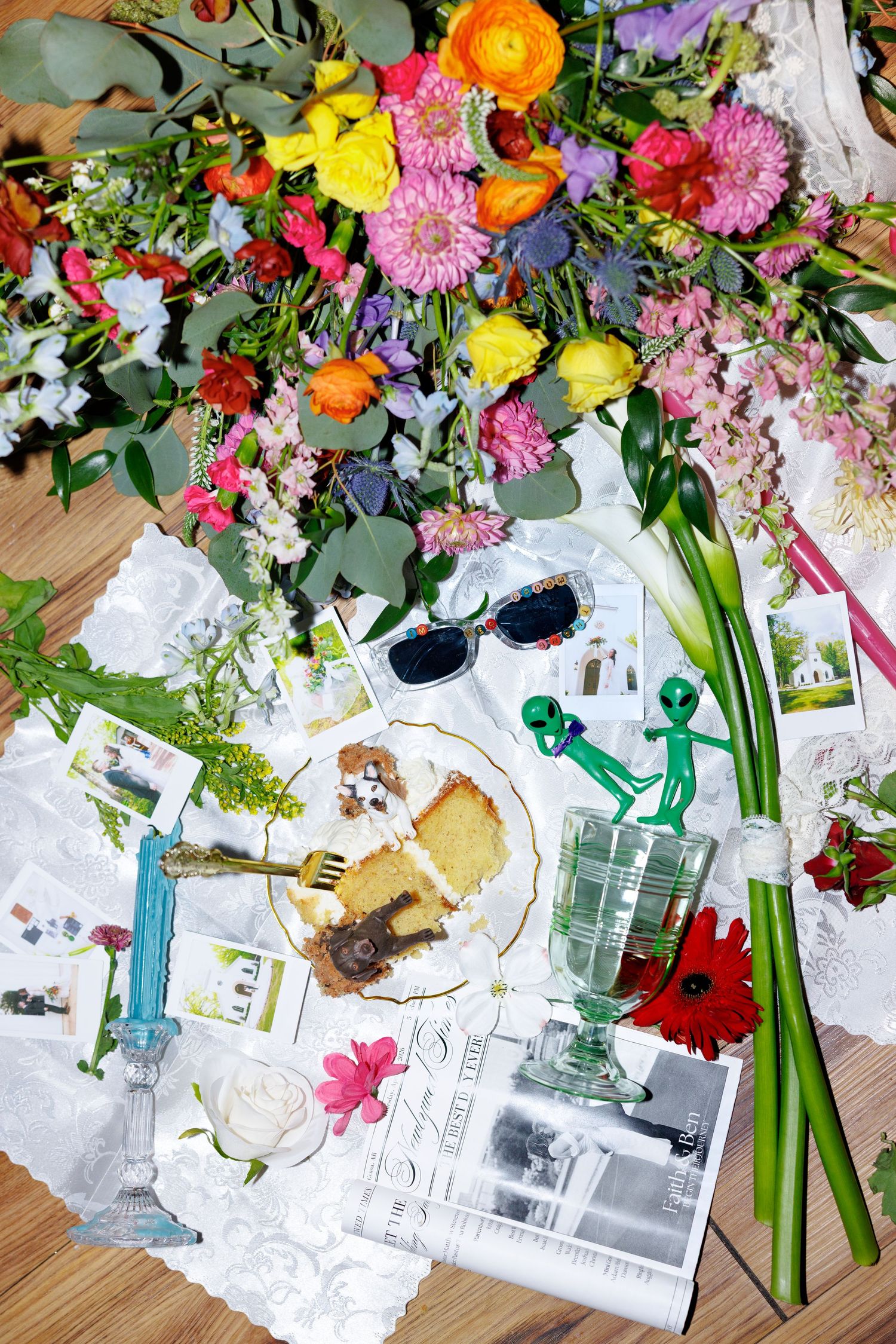 Colorful floral bouquet surrounded by cake, sunglasses, alien figurines, candles, and scattered photos on a table.