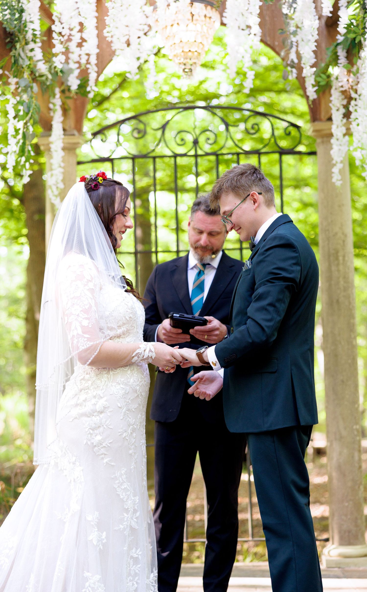 Bride and groom exchange rings at outdoor wedding altar with officiant beneath ornate iron gate and florals.