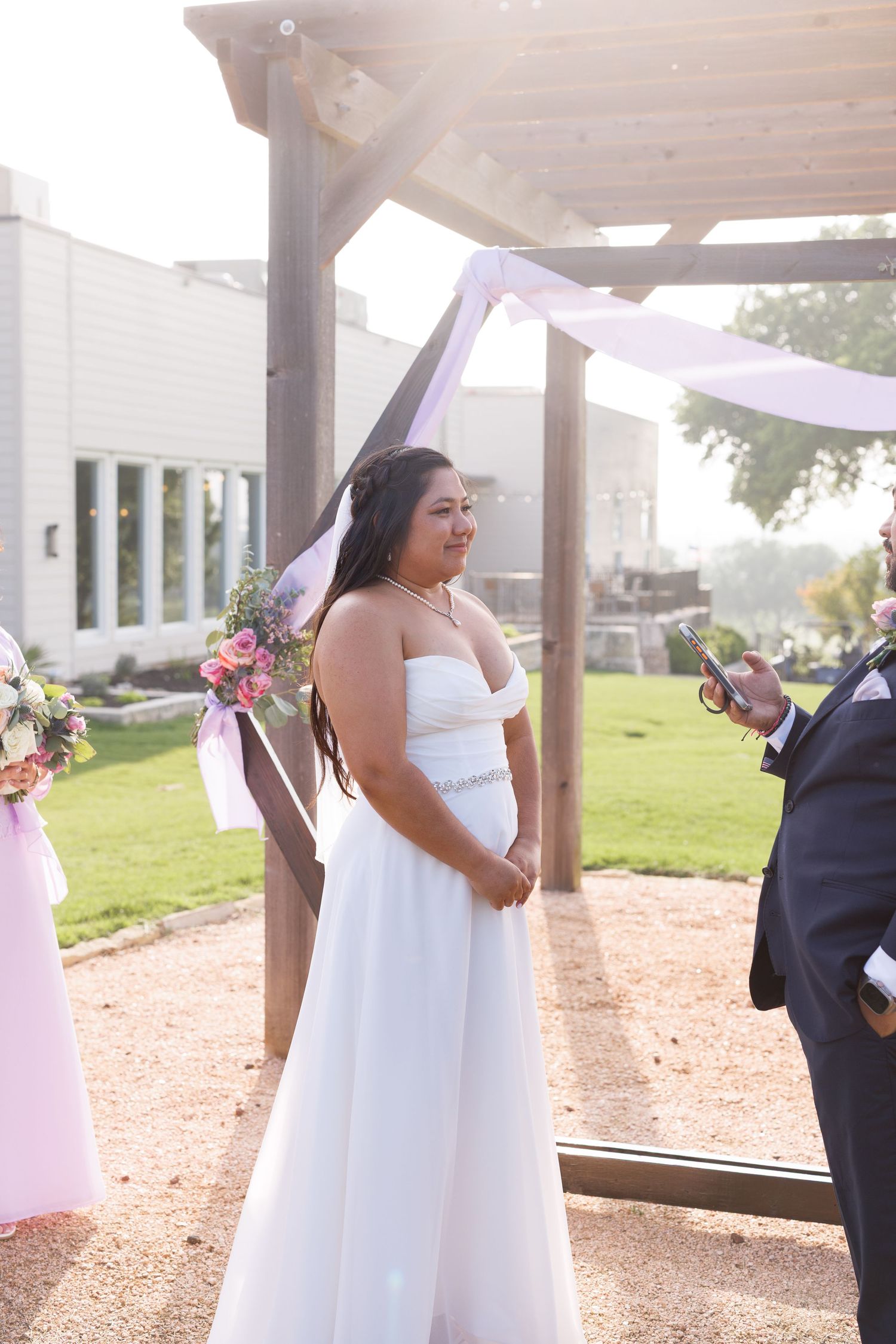 Bride in white strapless gown holding bouquet at outdoor wedding ceremony under wooden pergola.