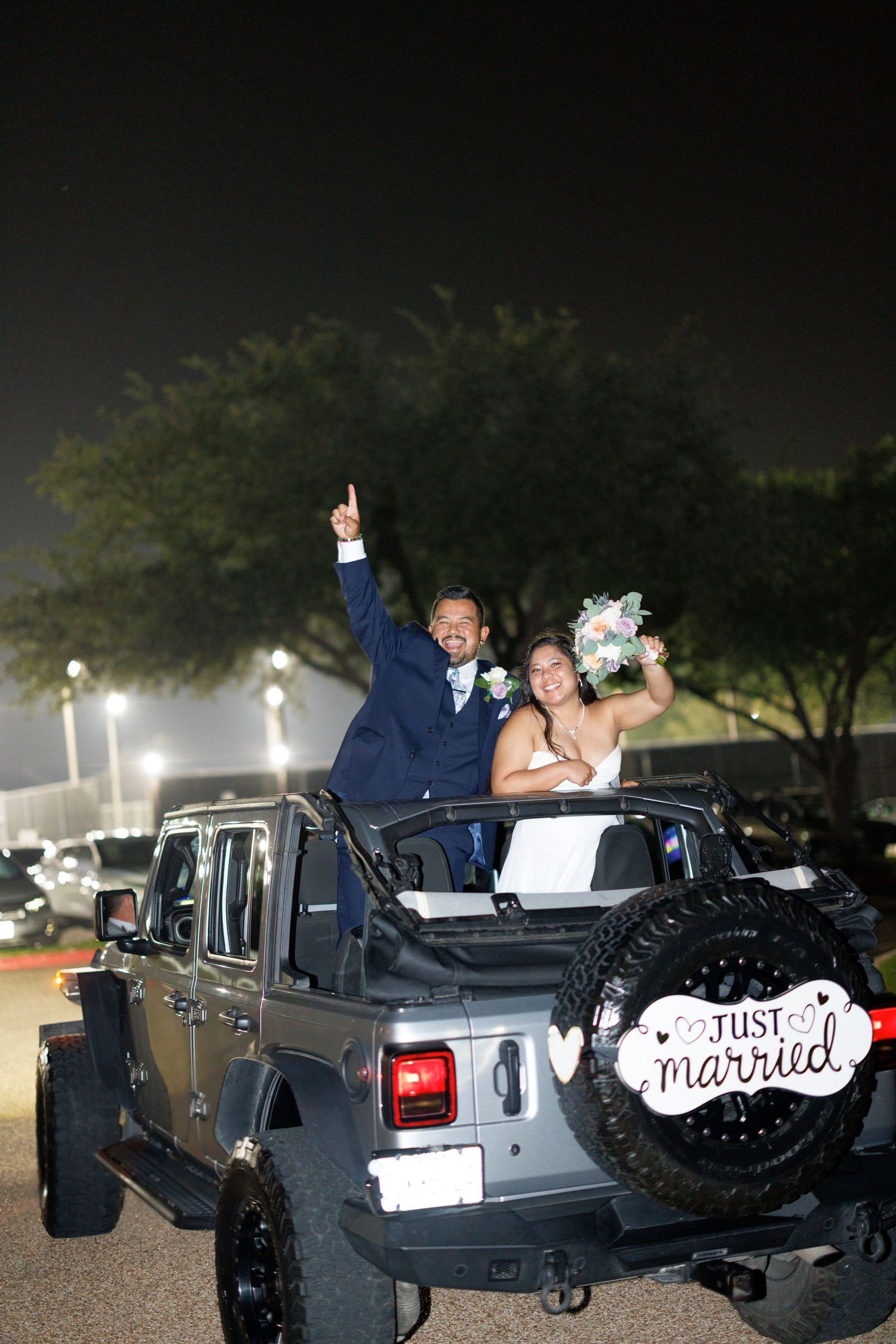 Newlyweds celebrate in a gray Jeep Wrangler with a Just Married sign at night.