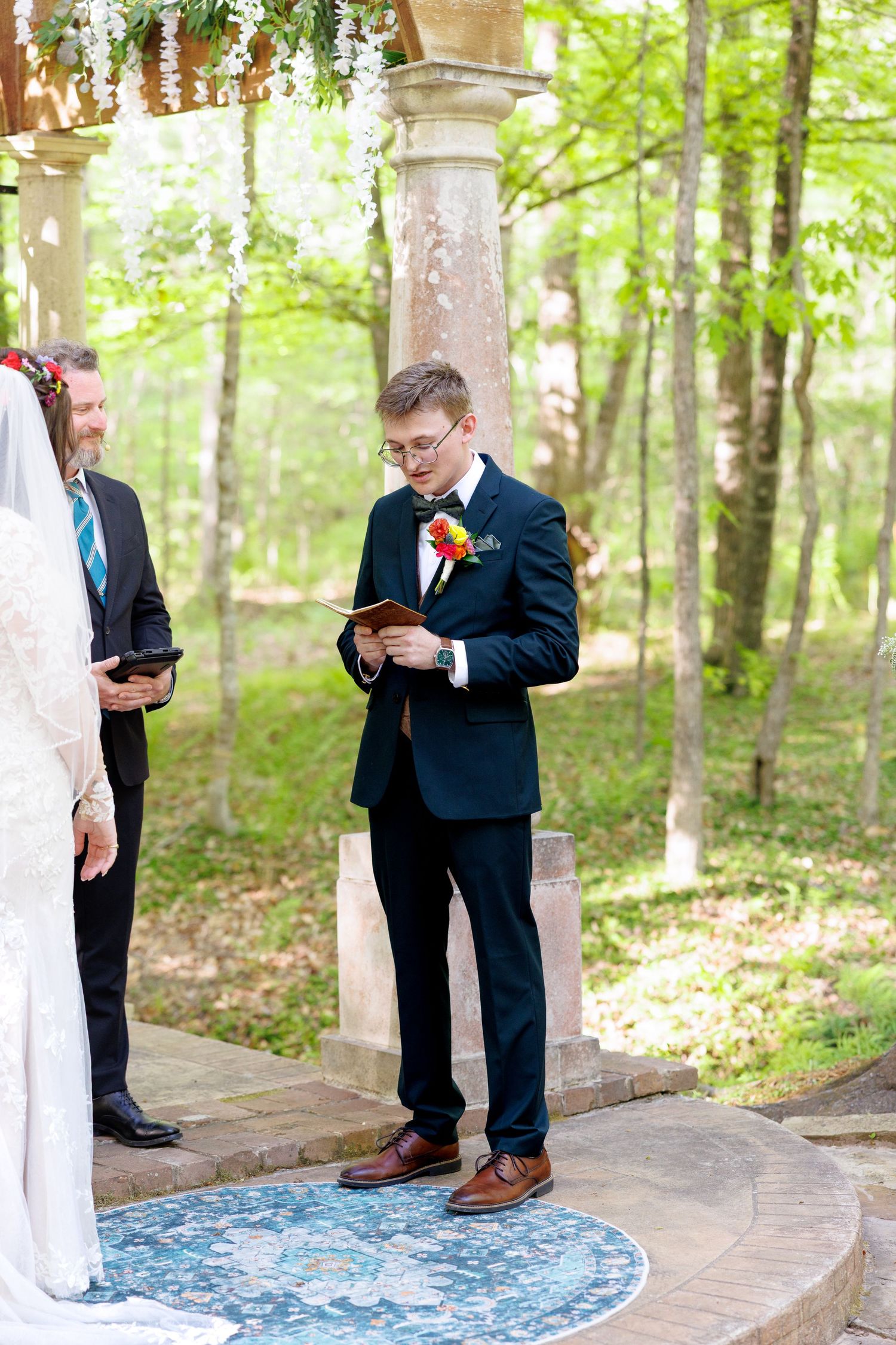 Groom in black suit reads vows from book at outdoor woodland wedding ceremony beneath floral arch.