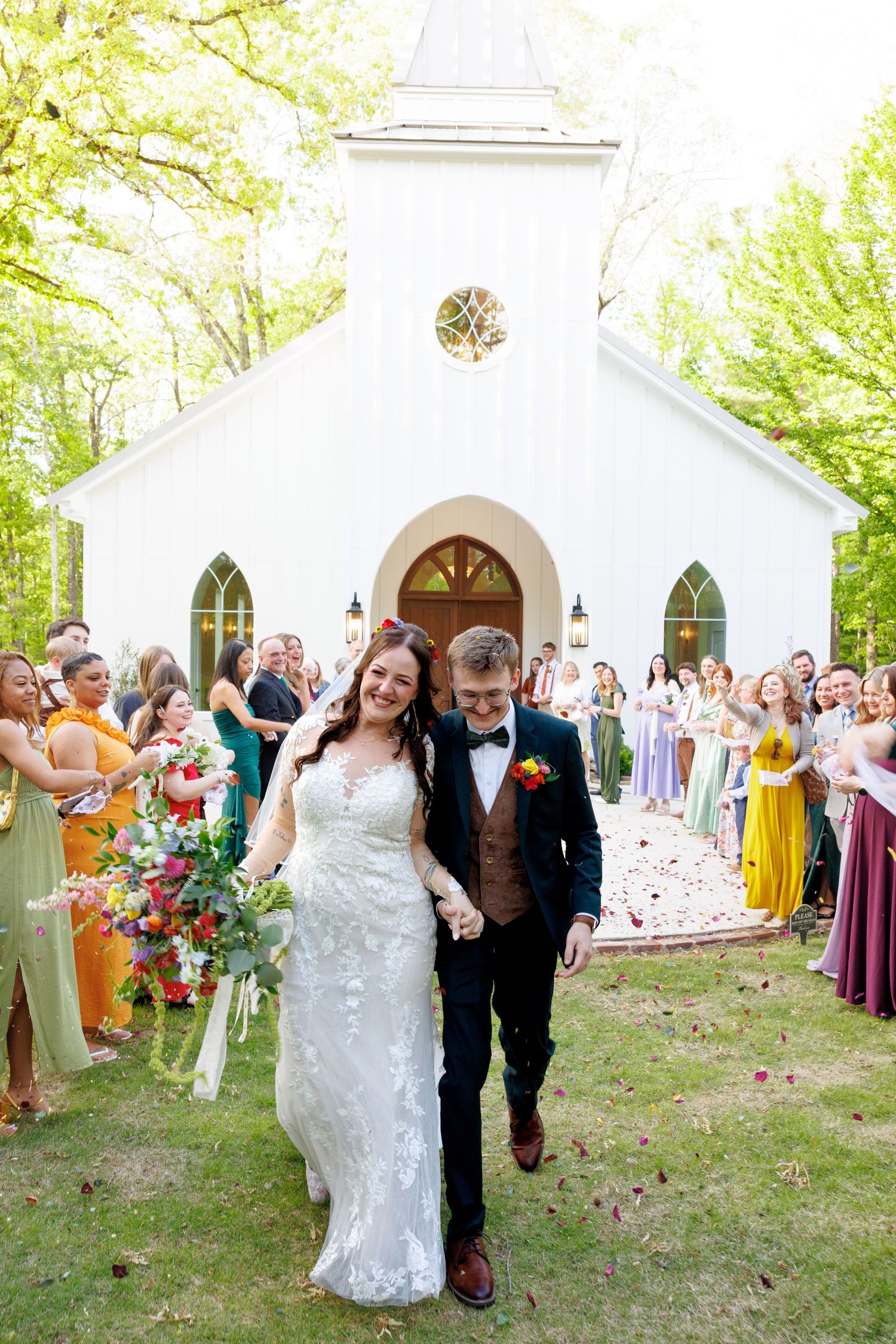 Joyful newlyweds exit a white chapel as guests celebrate with flower petals in a sunny outdoor setting.