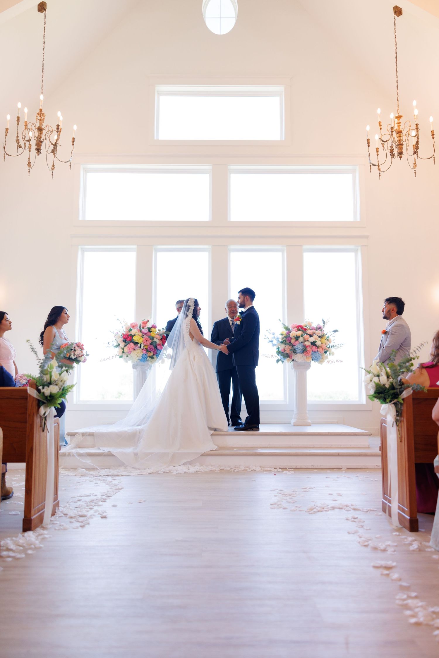 Bright white chapel interior with wooden pews and elegant chandeliers during wedding ceremony.