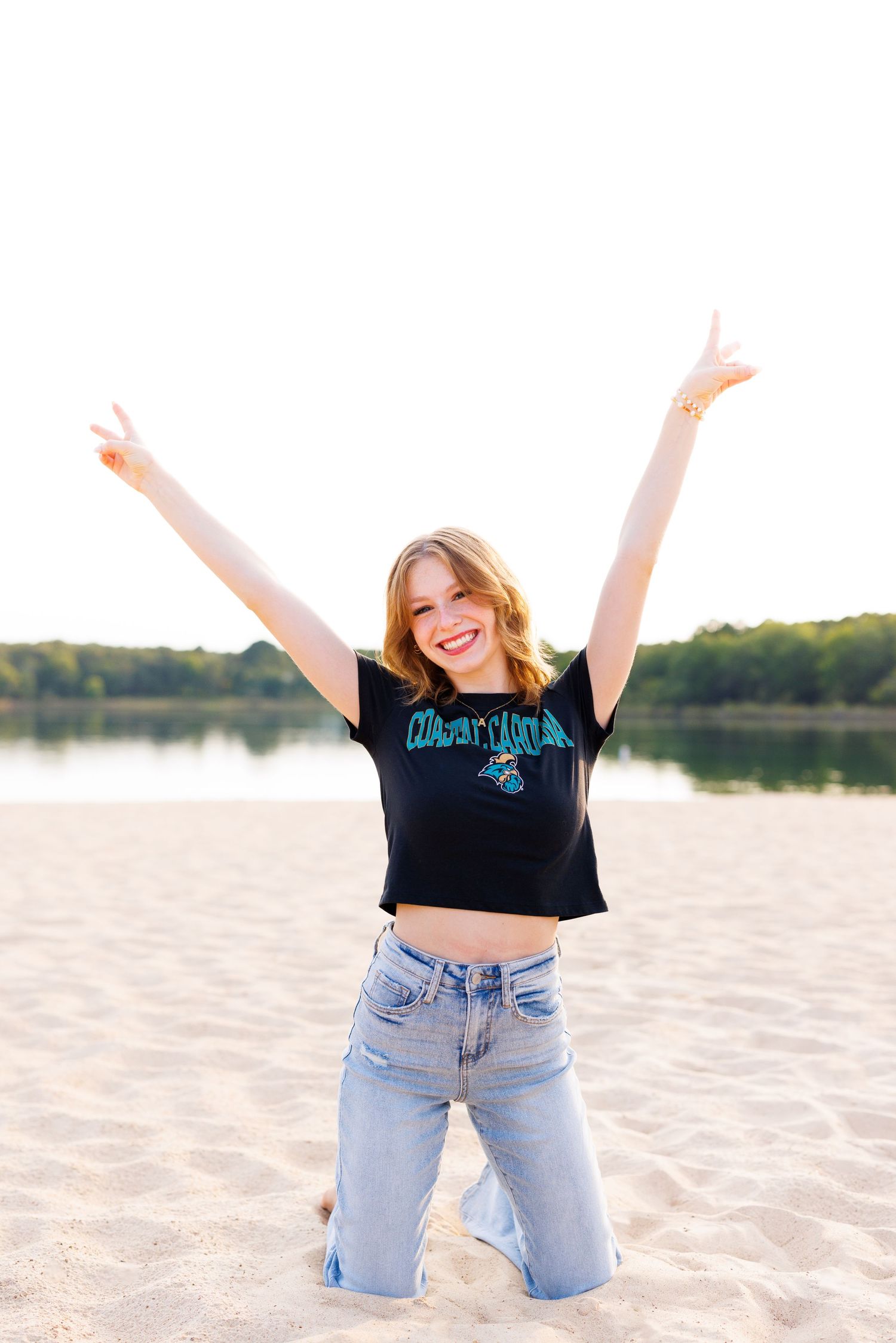 Person in black crop top and jeans jumps joyfully on beach with arms raised.