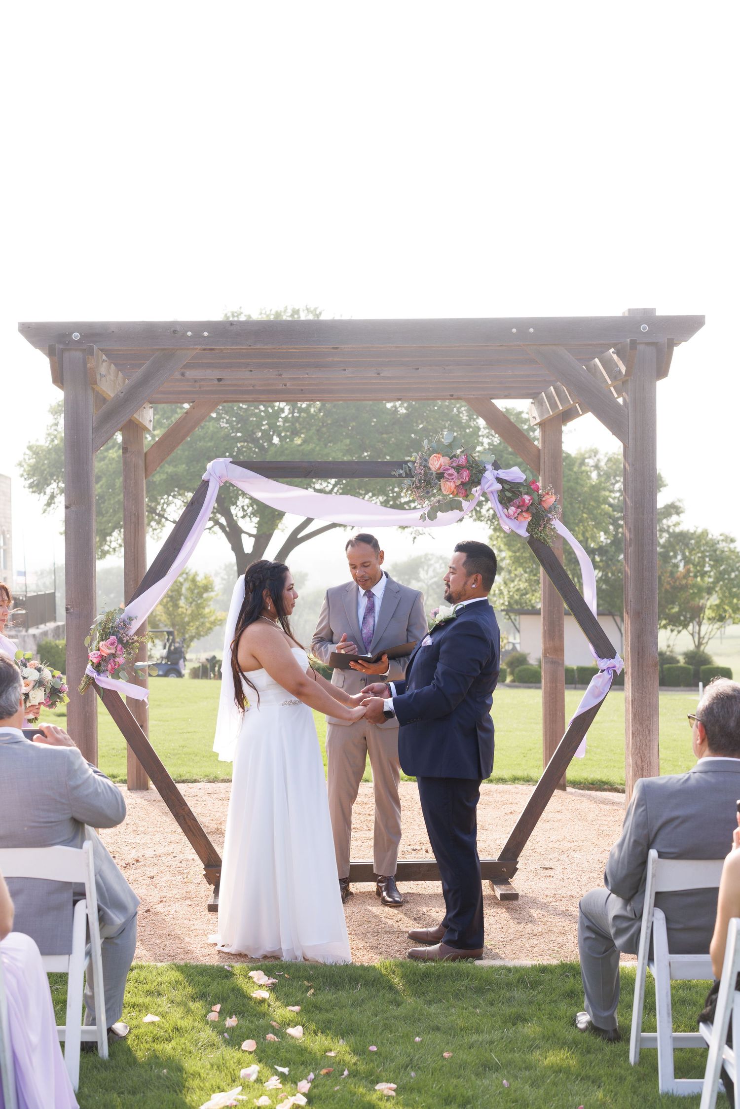 Bride and groom exchange vows under rustic wooden pergola decorated with flowers at outdoor wedding ceremony.