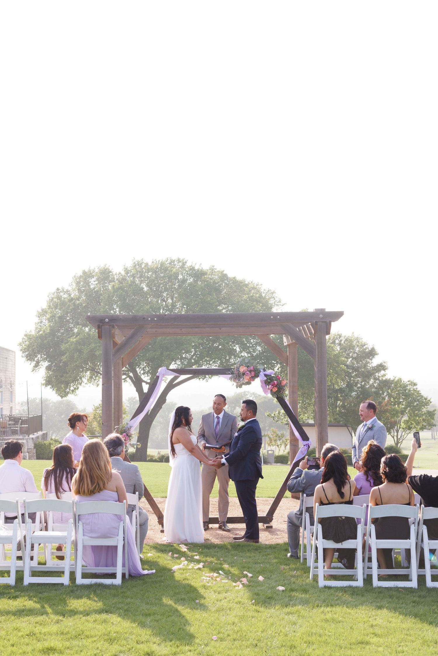 Outdoor wedding ceremony with couple at wooden arch, white chairs, and lush green lawn under bright sky.
