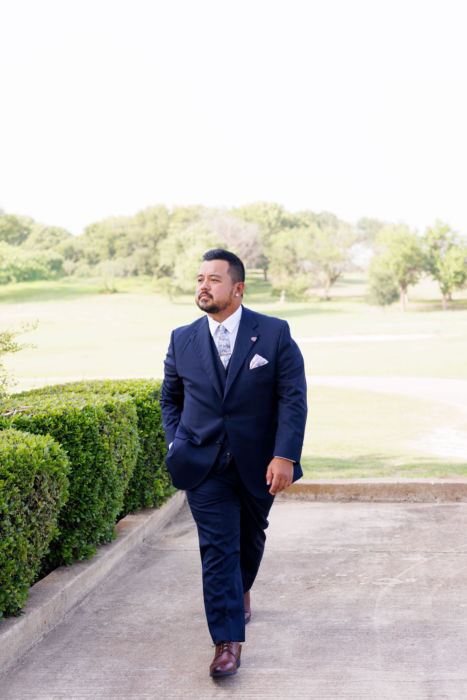 Groom walking confidently in a navy blue three-piece suit with white pocket square on a path beside manicured hedges.