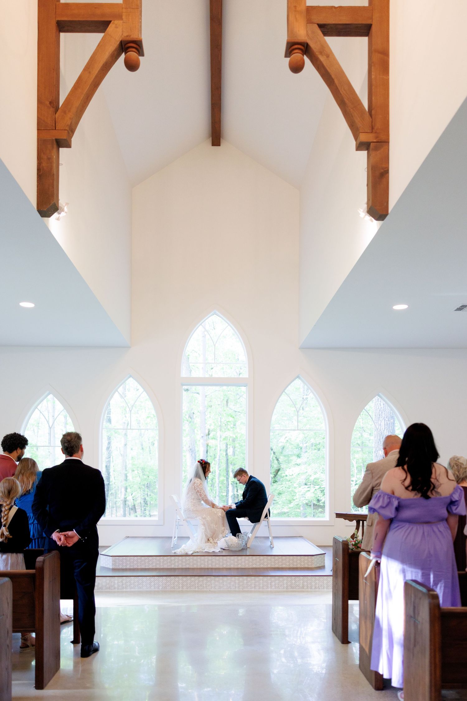 Wedding ceremony inside white chapel with arched windows, couple at altar with guests seated in wooden pews.