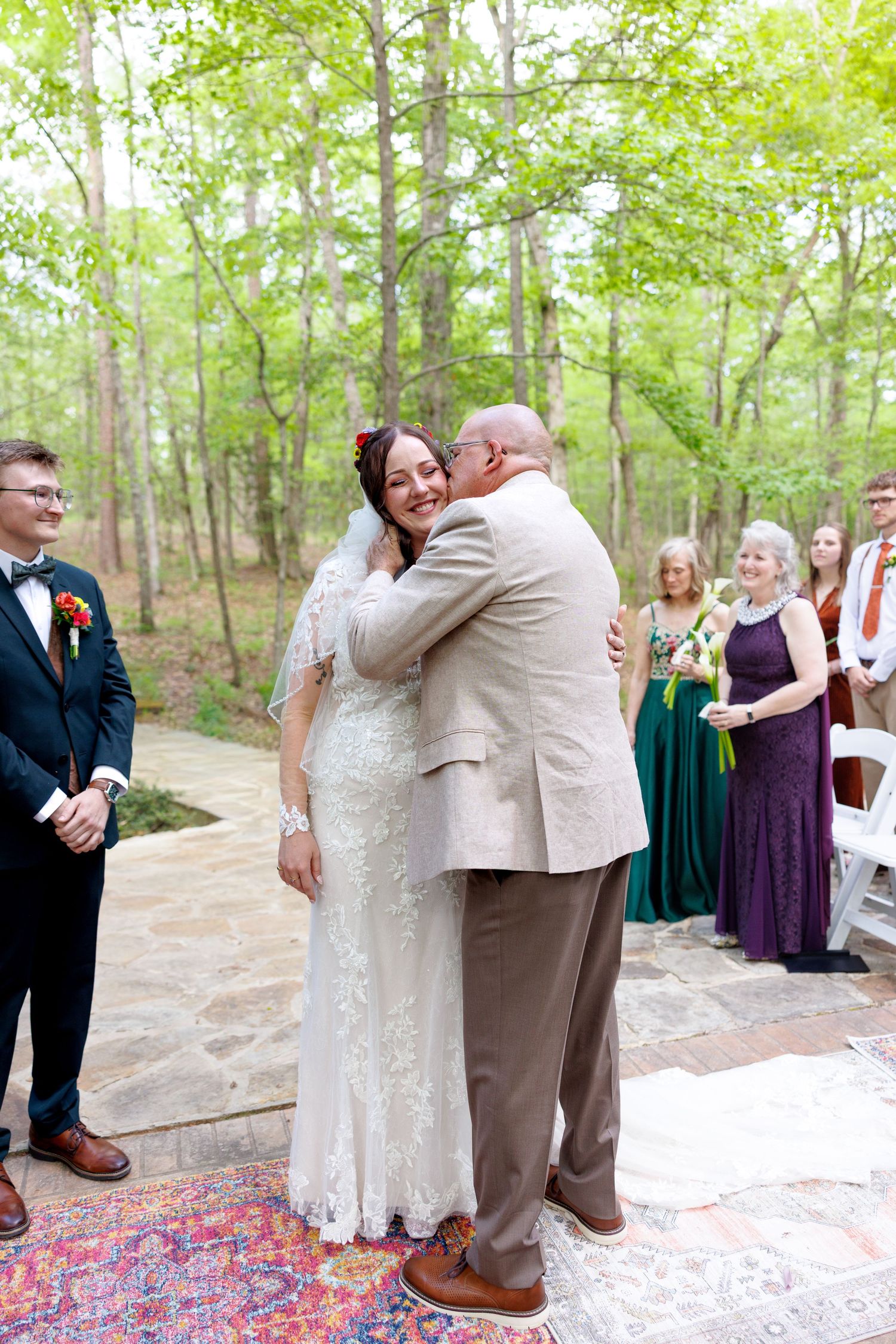 Smiling bride in lace dress embraces escort at outdoor wedding ceremony with bridesmaids in green gowns.