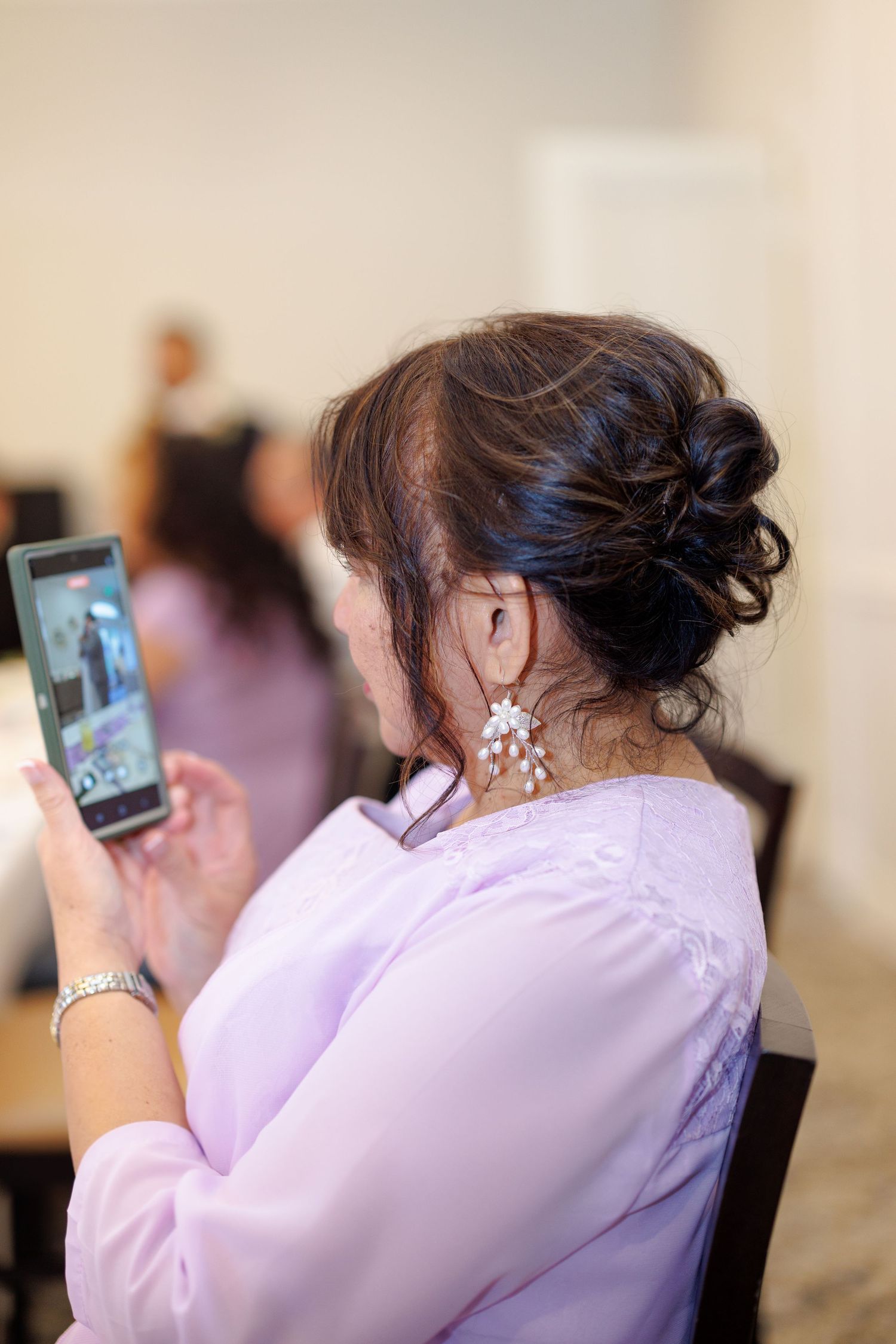 Woman with elegant updo and chandelier earrings photographing on smartphone at a formal indoor event.