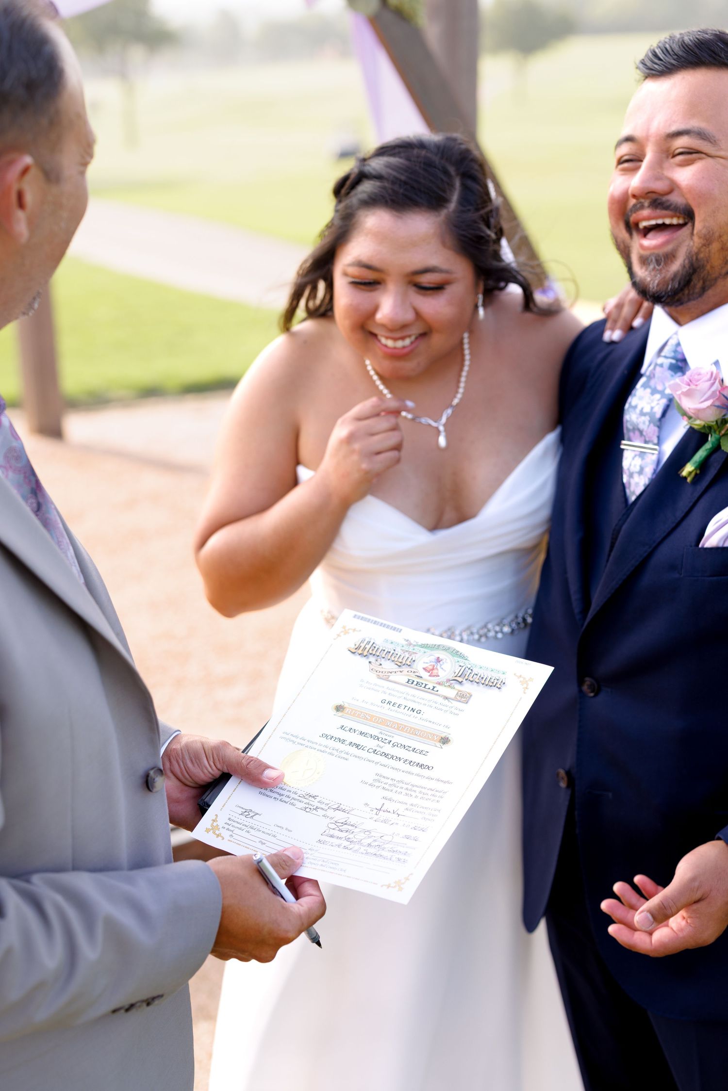 Laughing bride and groom sign marriage certificate with officiant during joyful outdoor wedding ceremony.