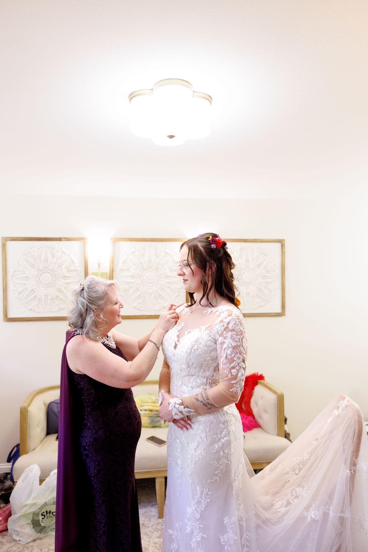 Older woman in purple dress helps bride in white lace gown with necklace in elegant bridal suite.
