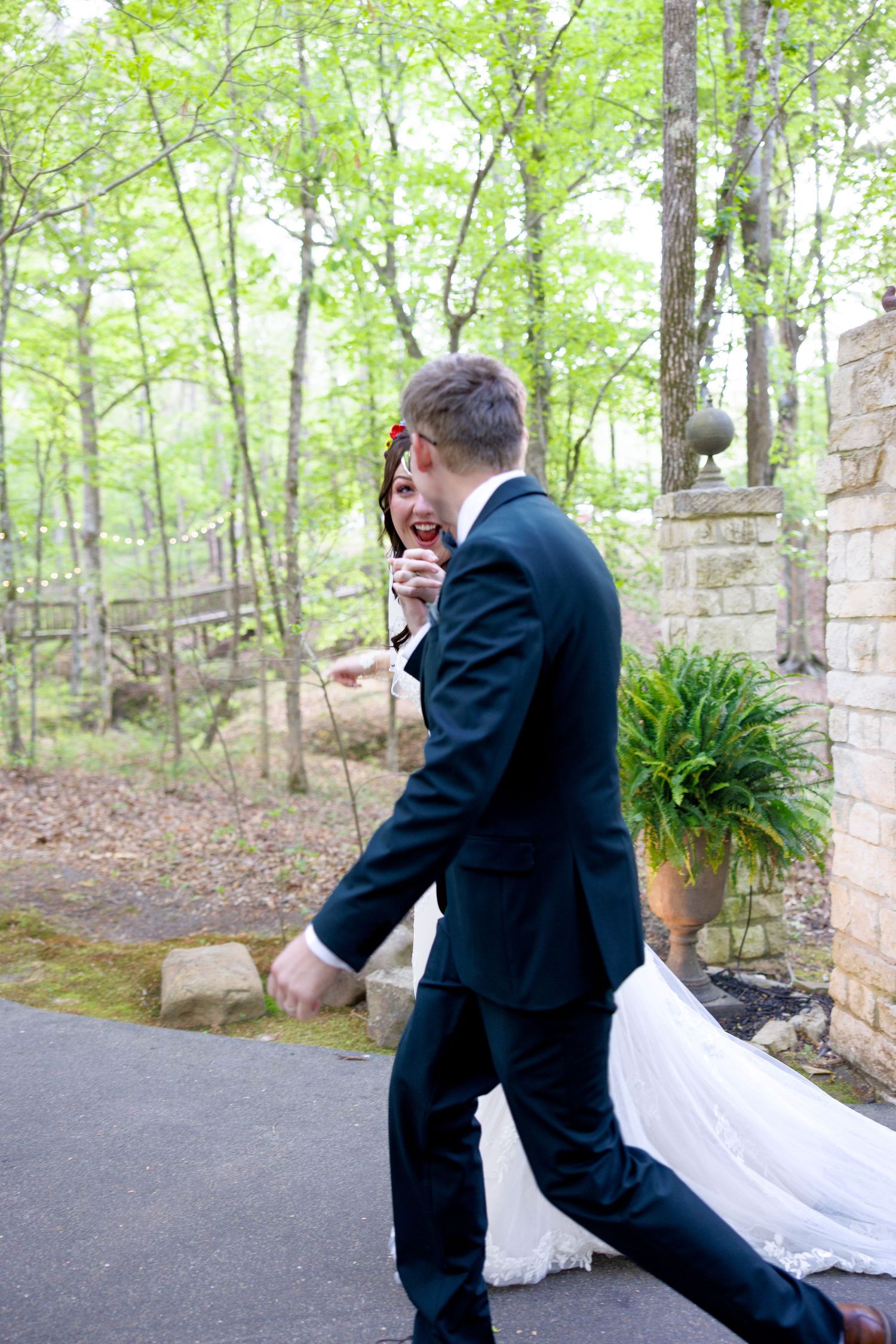 A joyful bride and groom walk together outdoors, laughing, surrounded by lush green trees at their wedding venue.
