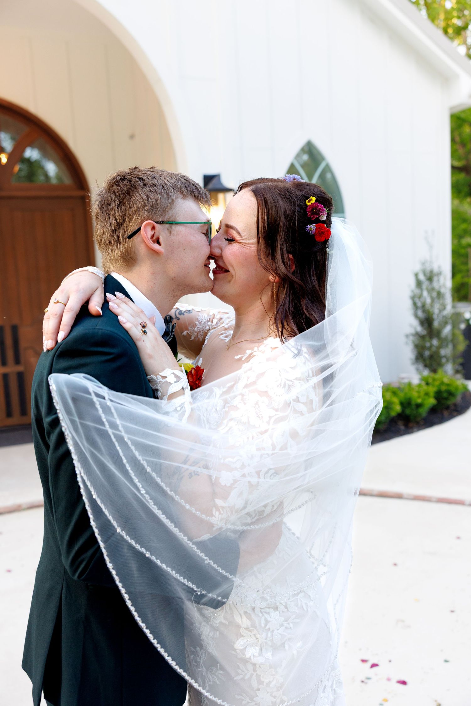 Bride and groom share a romantic kiss outdoors, bride's veil flowing around them near a white chapel.
