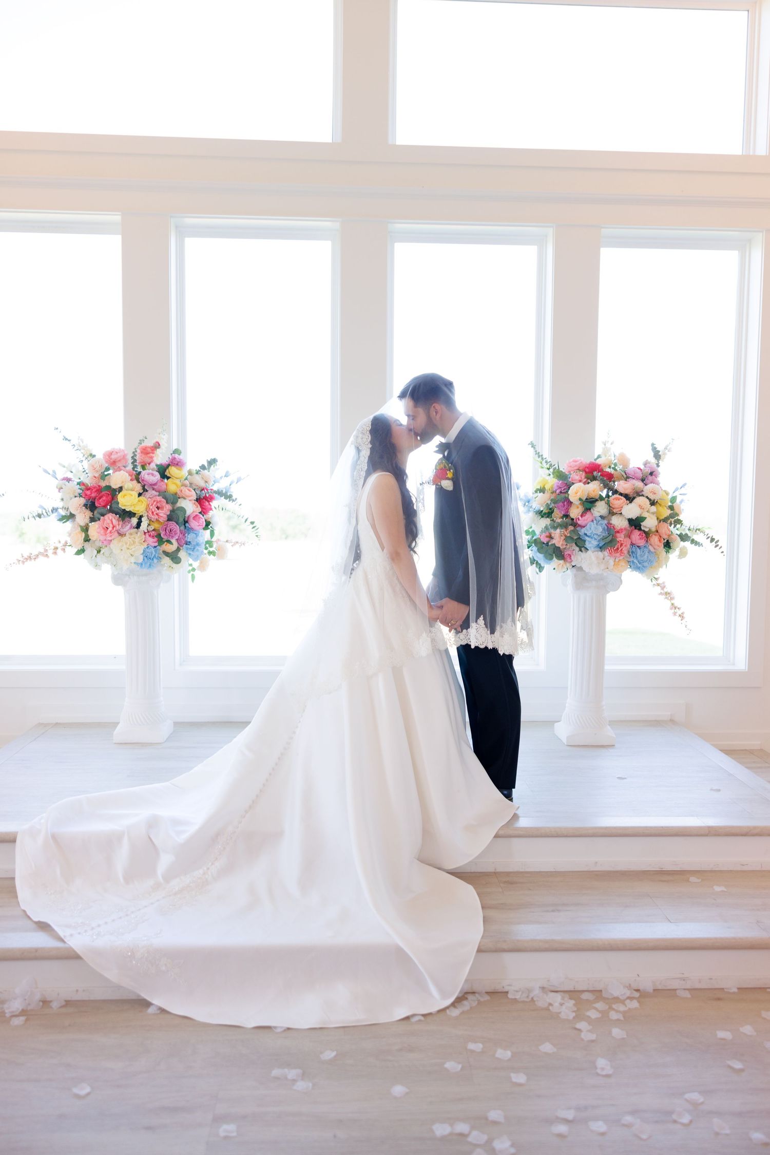 Romantic wedding couple shares an intimate moment in a bright white room decorated with colorful floral arrangements.