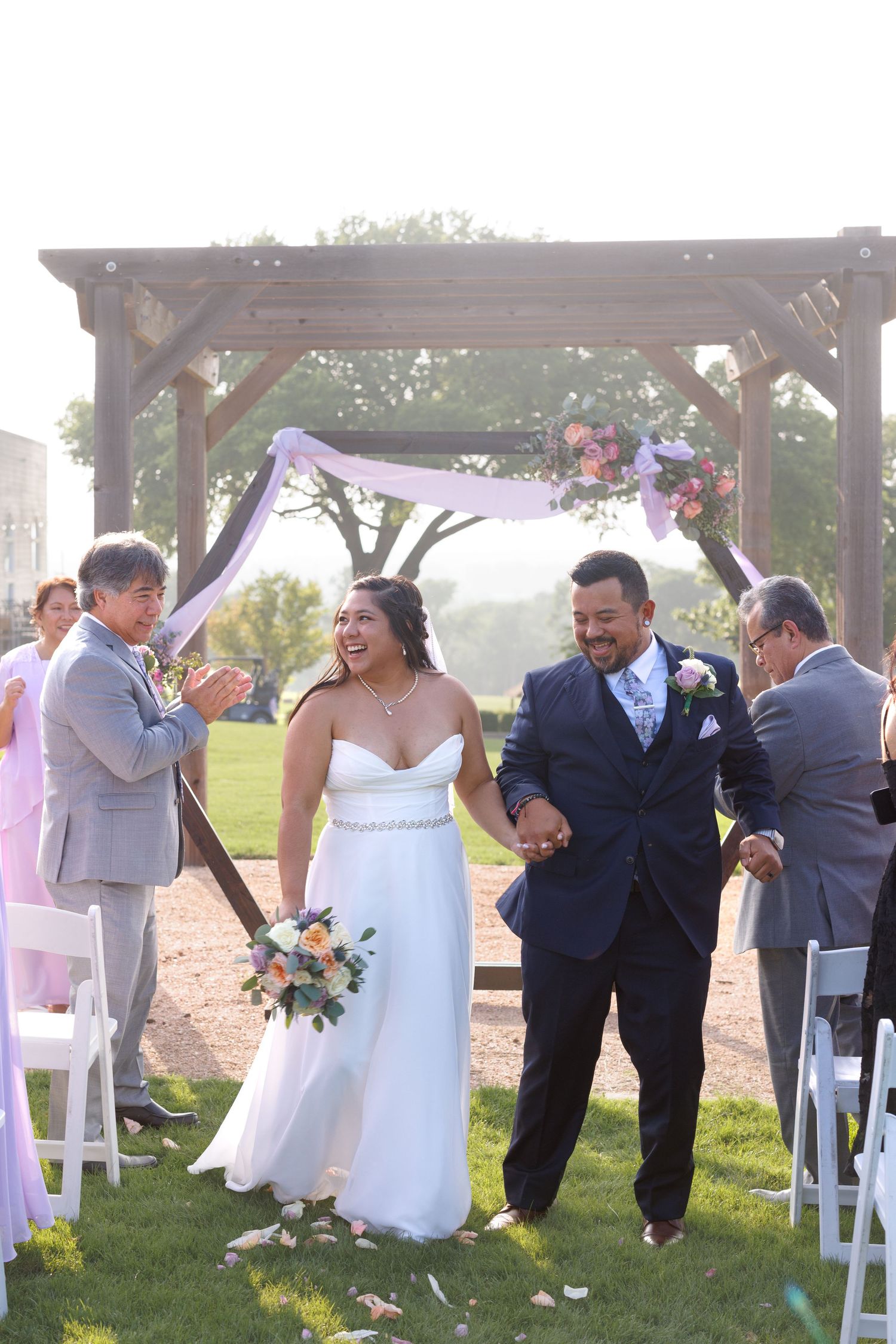 Newlyweds walk hand in hand down aisle after outdoor wedding ceremony under rustic wooden arch.
