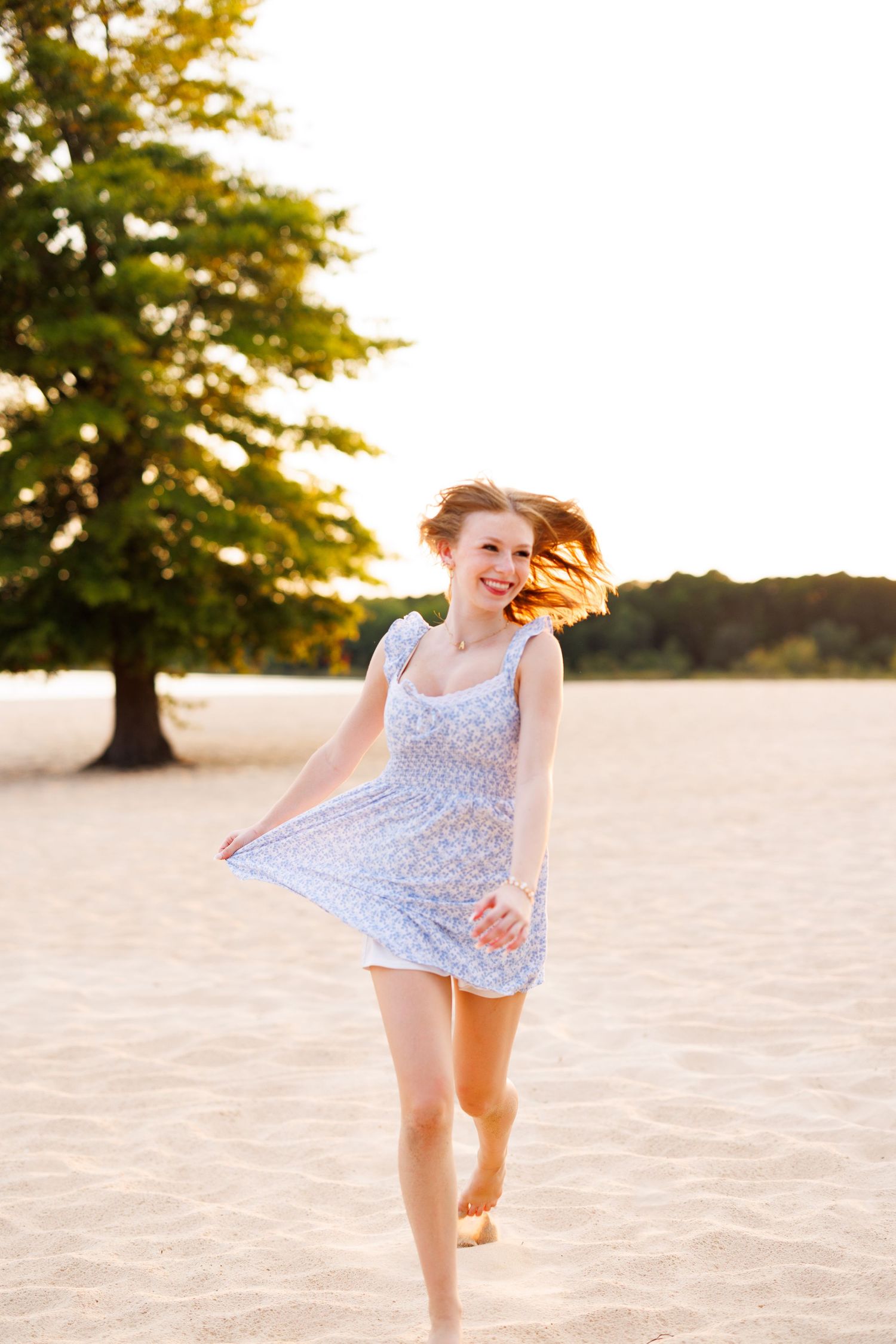 Carefree dance in floral blue dress on sandy beach with tree silhouette at sunset.