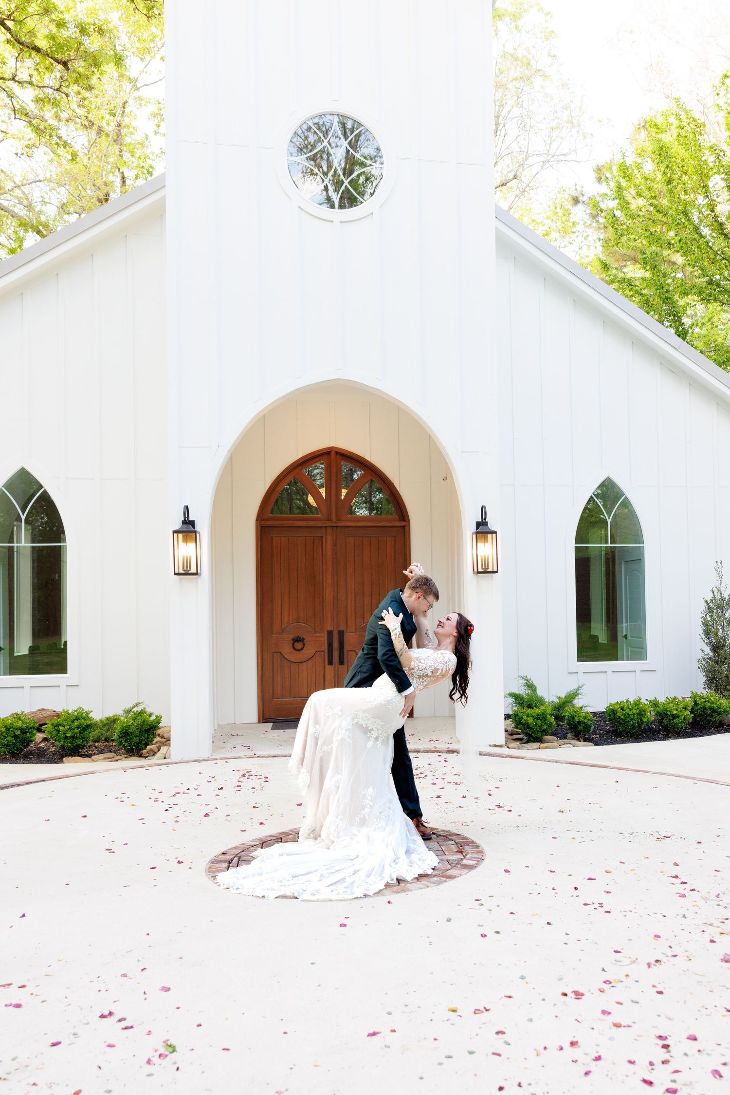Groom dips bride in a romantic kiss in front of a white chapel entrance with rose petals on the ground.