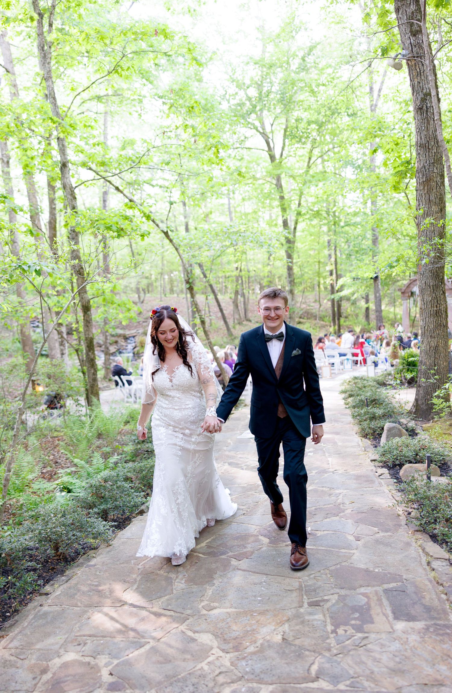 Bride and groom walk hand-in-hand down a stone path through a lush green forest after their outdoor wedding ceremony.