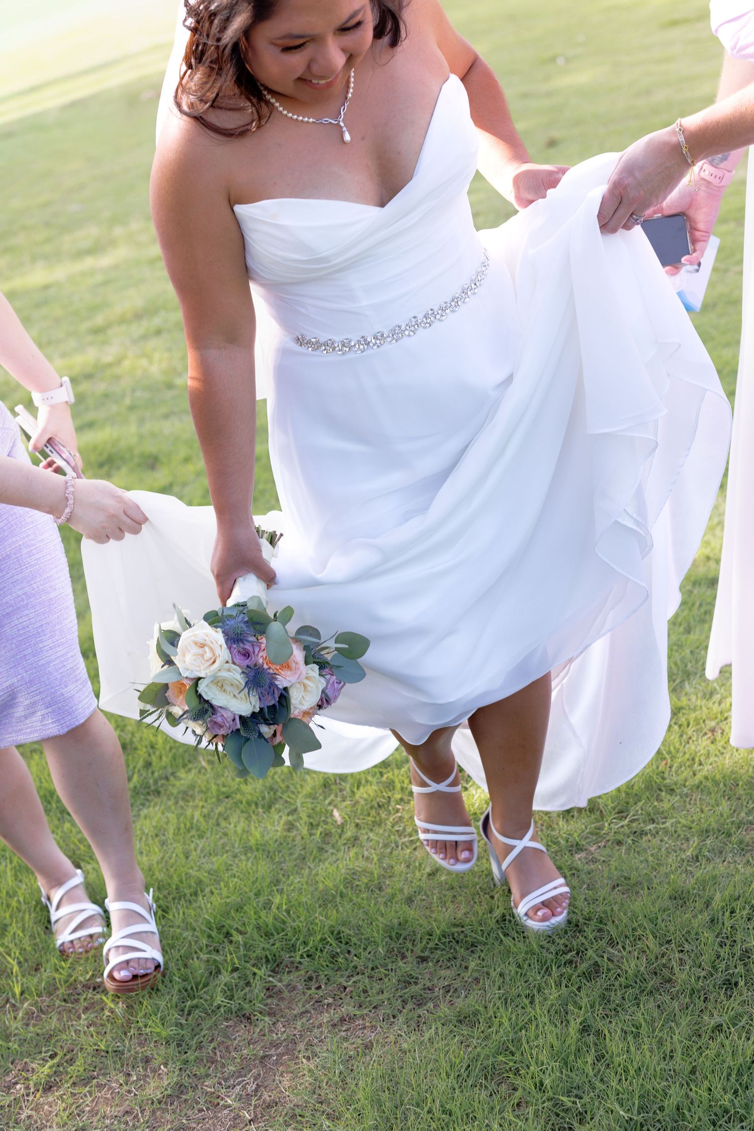 Bride in strapless white wedding gown walks across green grass while bridesmaids help carry her dress train.