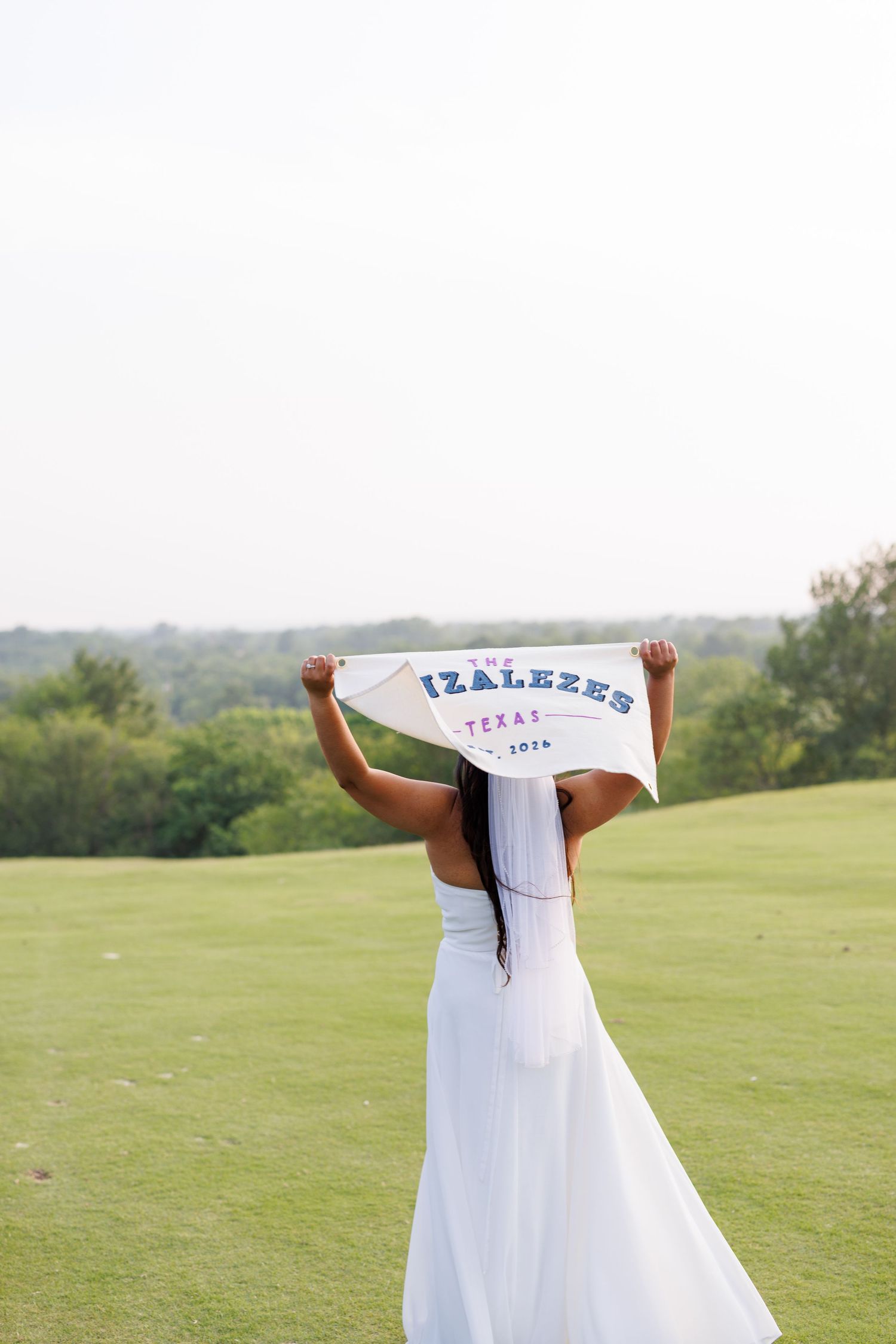 Bride in white gown holds The Izalezes Texas banner above her head on a lush green golf course.