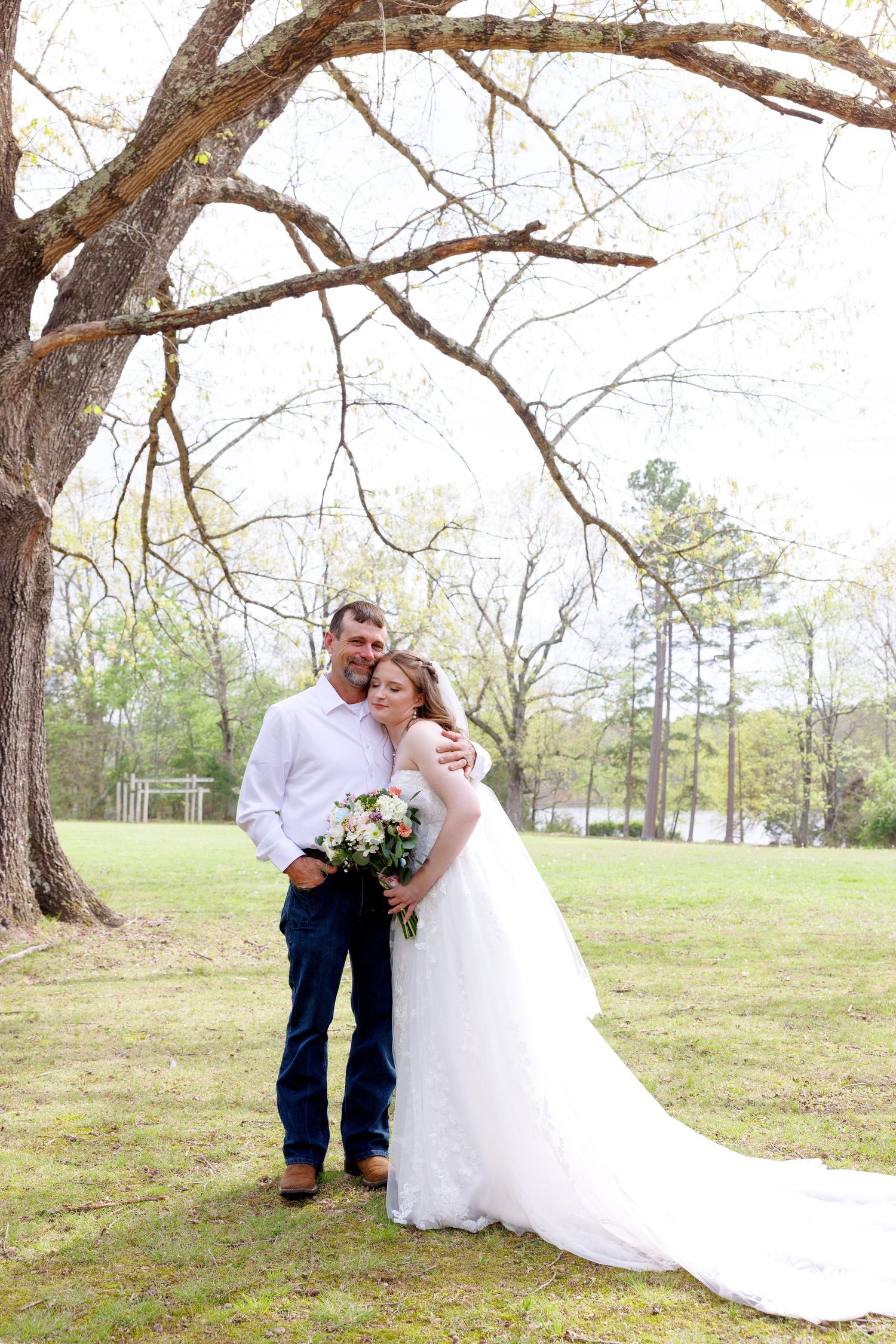 Bride in white gown holds bouquet while groom in jeans embraces her under a large oak tree in a green outdoor setting.