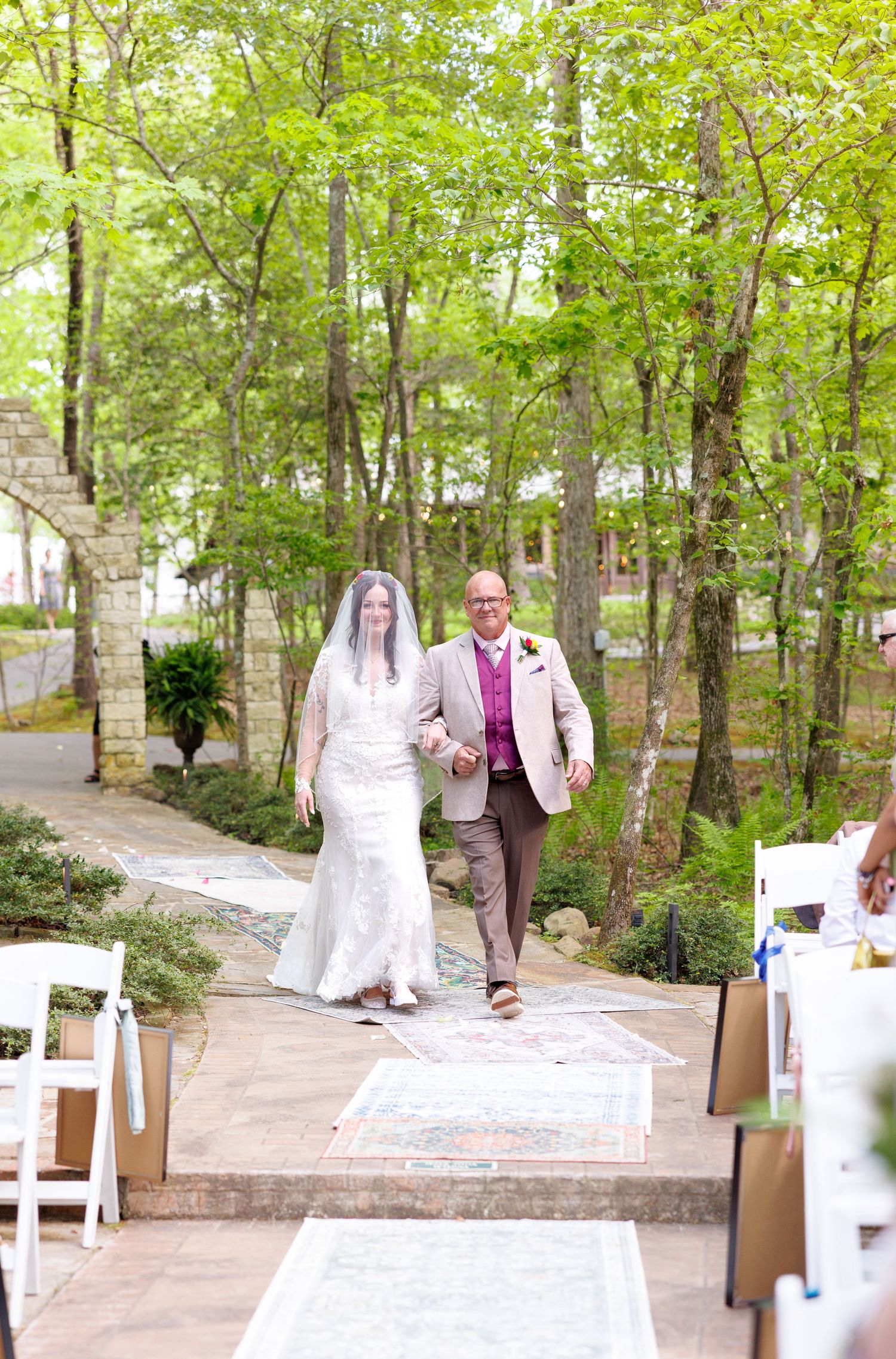 Bride and groom walking down outdoor wedding aisle surrounded by lush green trees.