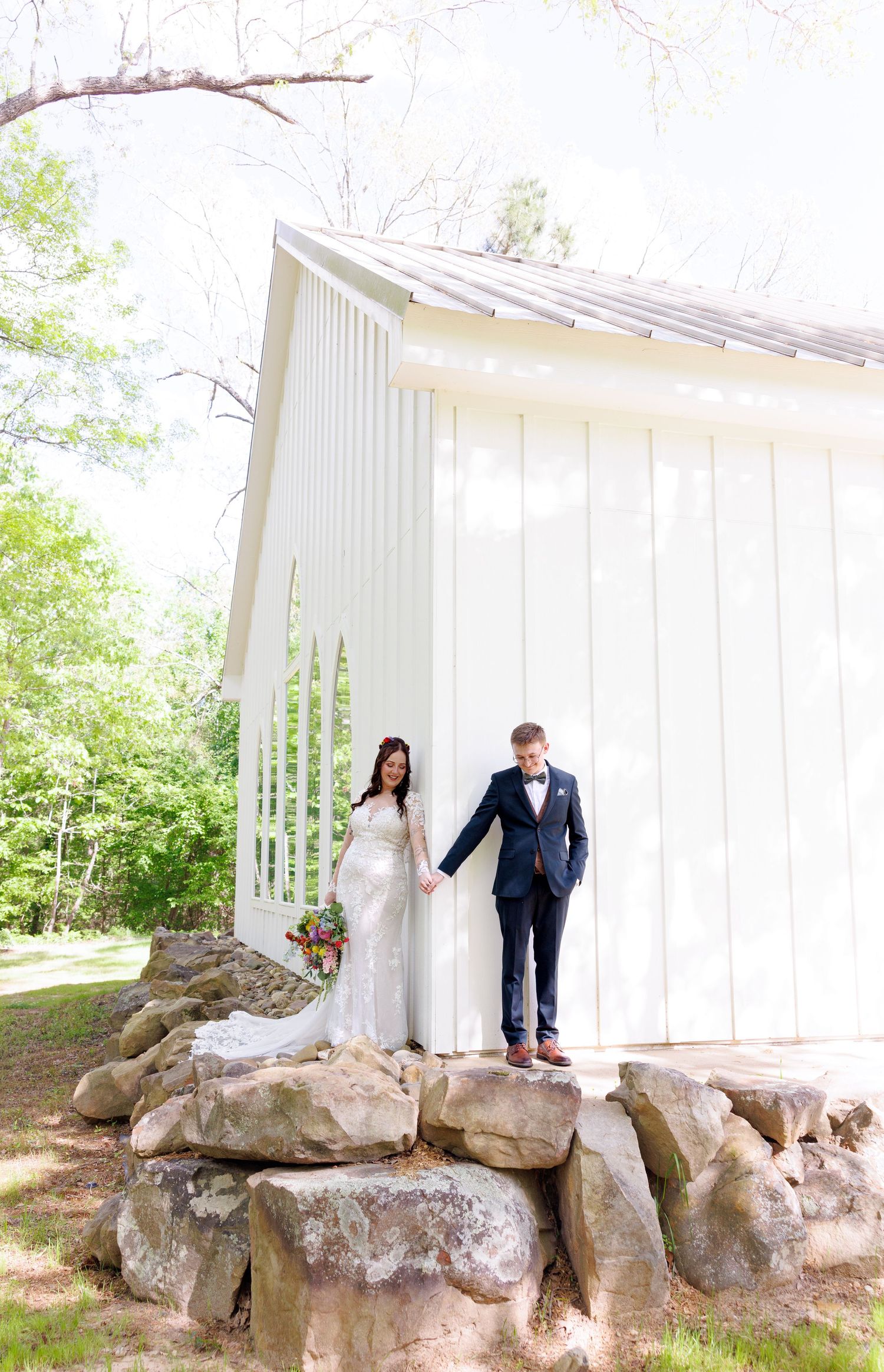 Bride and groom holding hands on rocks beside white chapel surrounded by lush green trees.