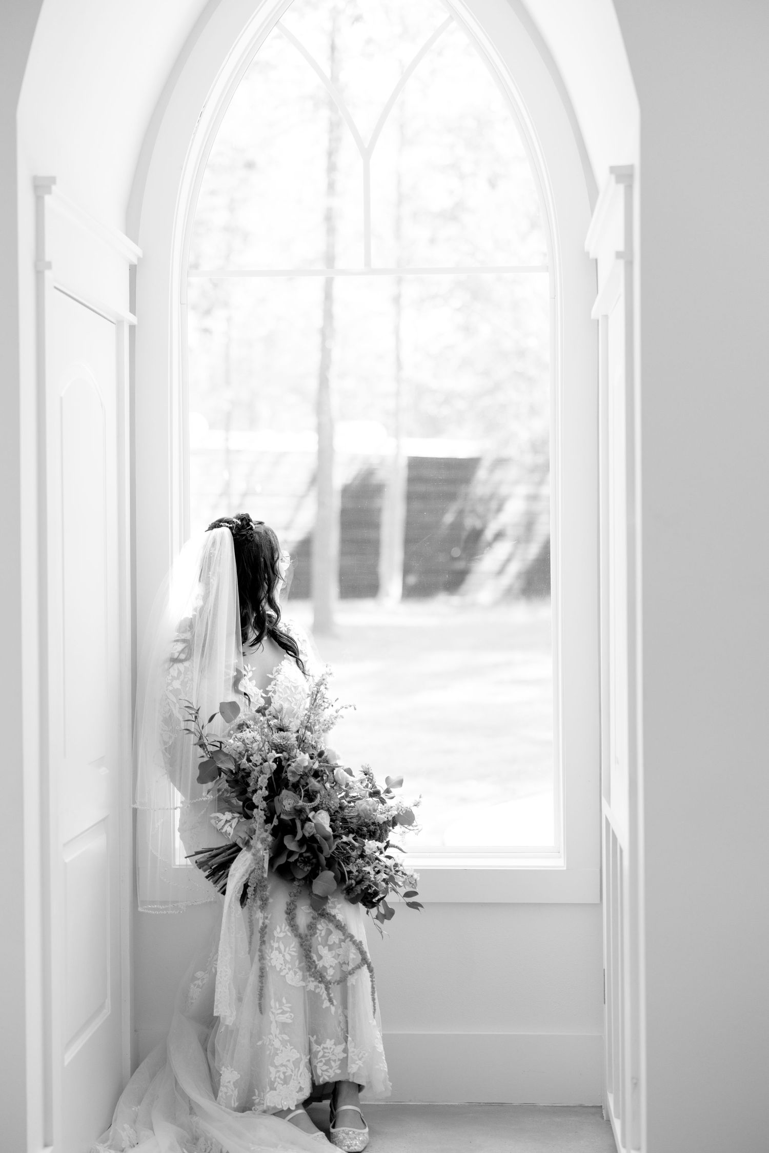 Black and white photo of bride in lace gown holding large bouquet standing before arched chapel window.