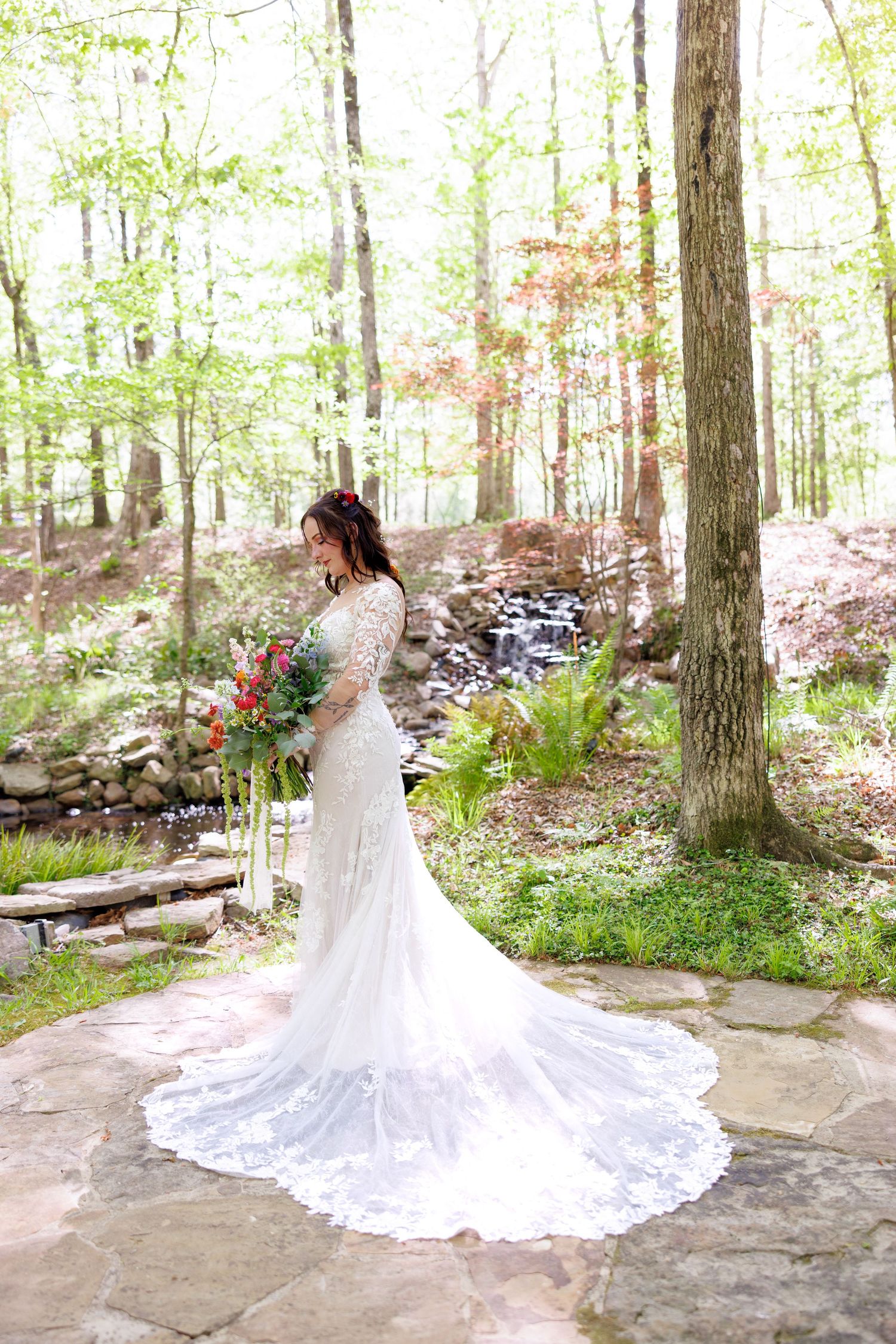 Bride in lace gown with long train poses in sunlit forest garden setting.