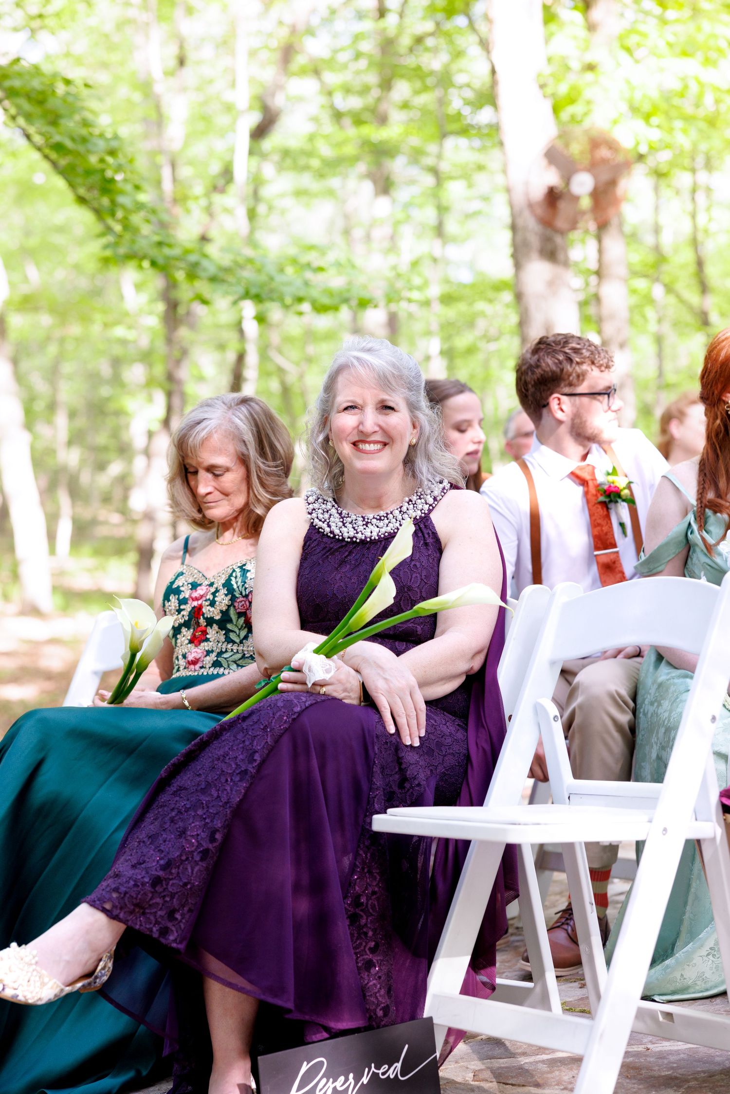 Woman in purple lace dress smiling at outdoor wedding ceremony seated in white chairs surrounded by guests in a forest.