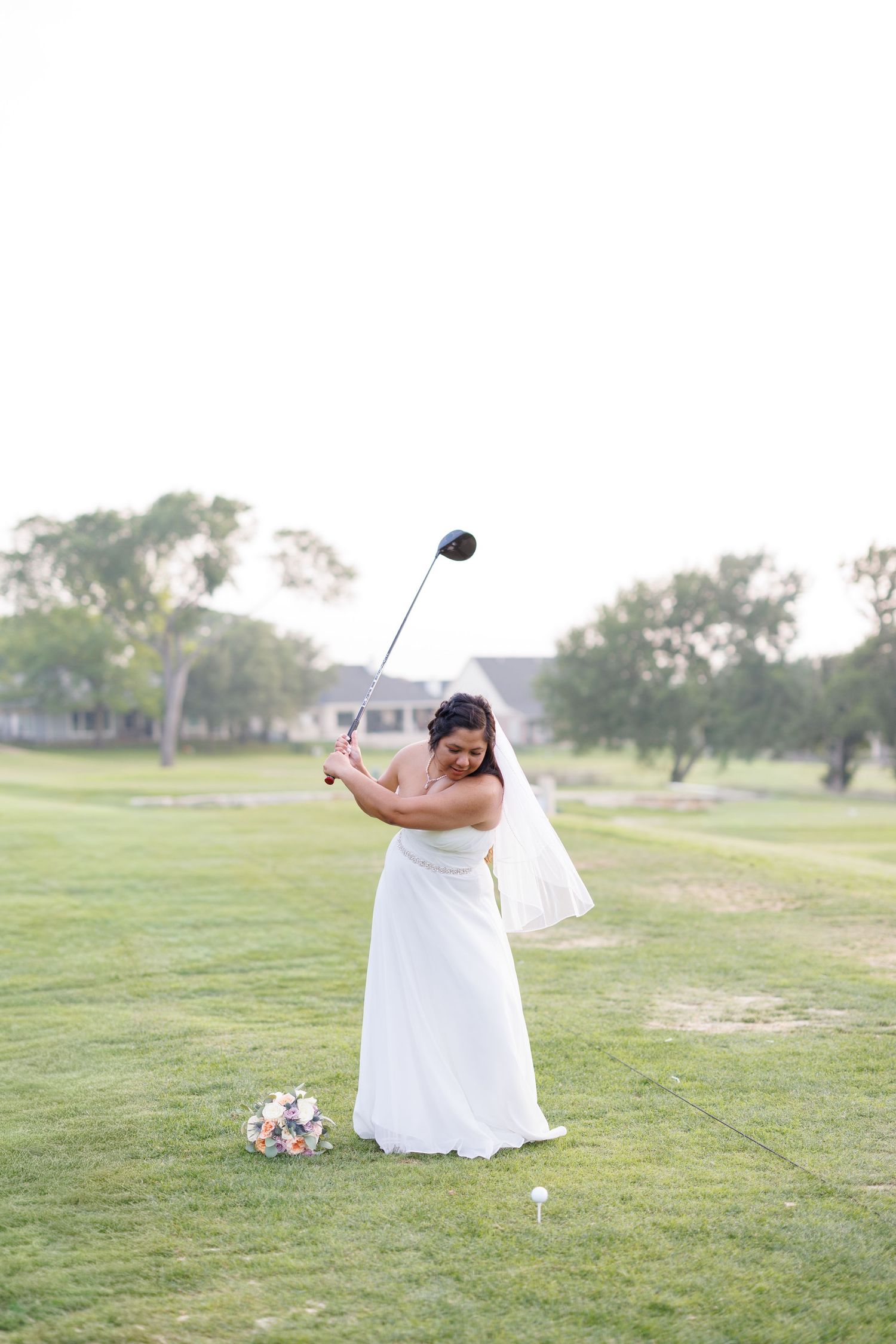Bride in white strapless gown and veil swings a golf club on a golf course with her bouquet nearby.