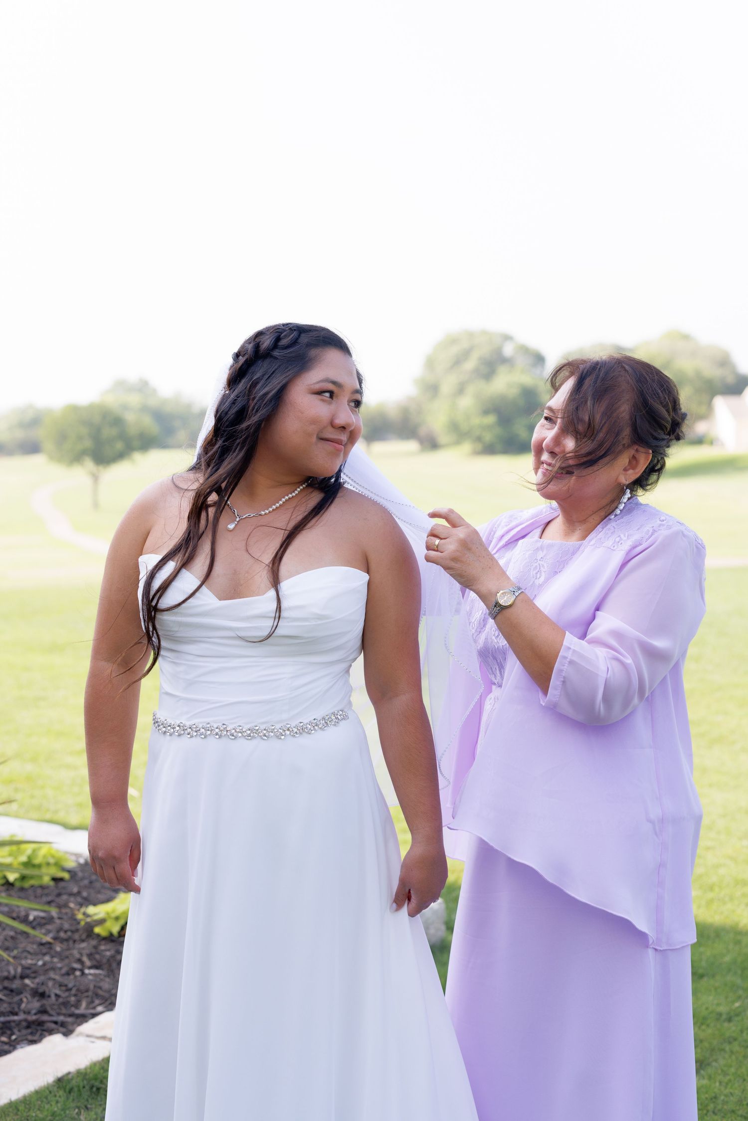 Bride in white strapless gown smiles as older woman in lavender adjusts her veil outdoors on a sunny day.
