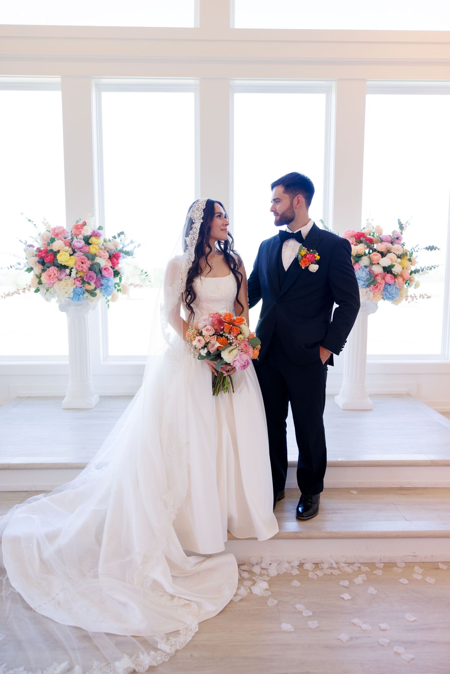 Wedding couple poses in bright white room with colorful floral arrangements and flowing dress train.