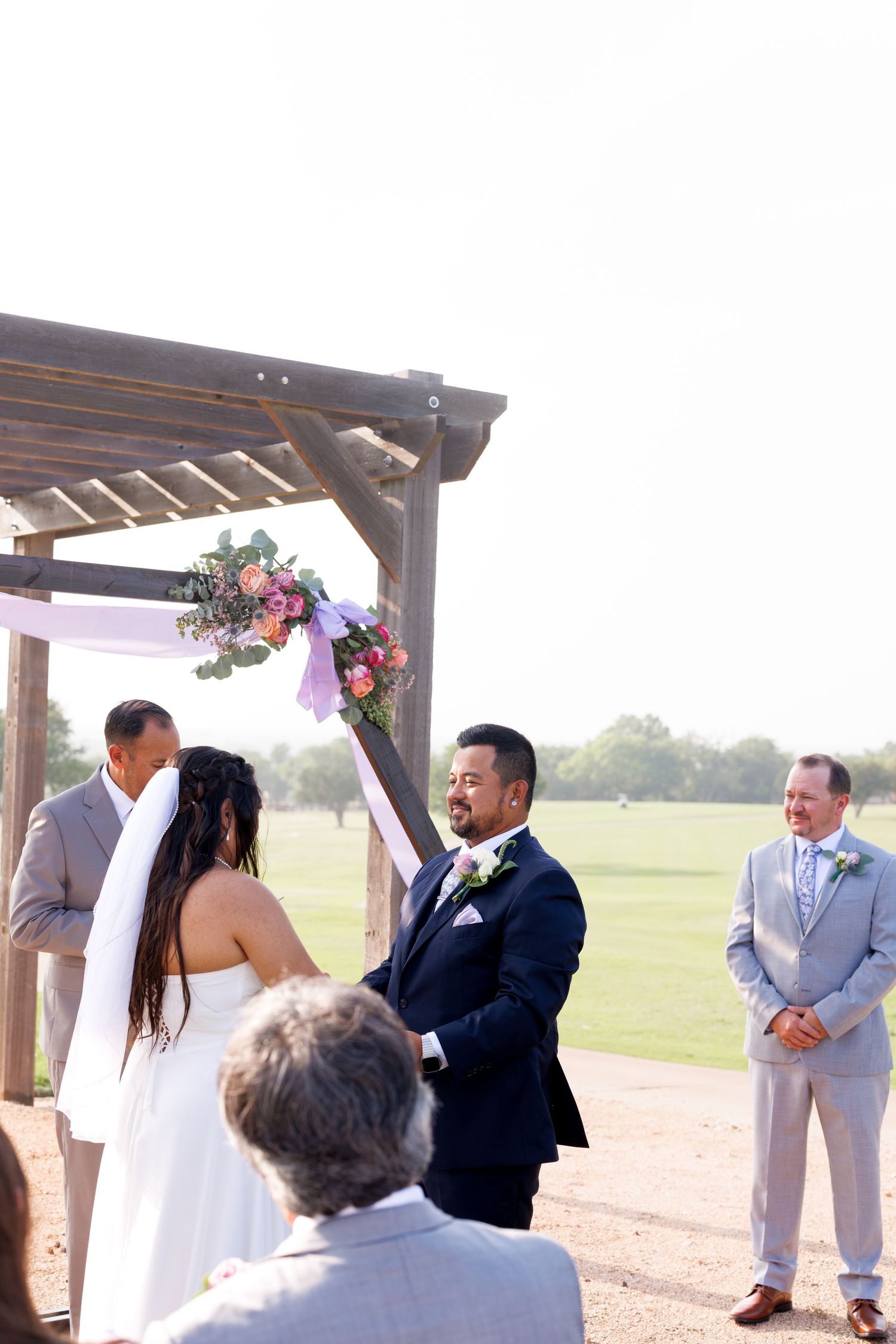 Groom in navy suit smiles at bride during outdoor wedding ceremony under rustic wooden arch with pink floral decor.