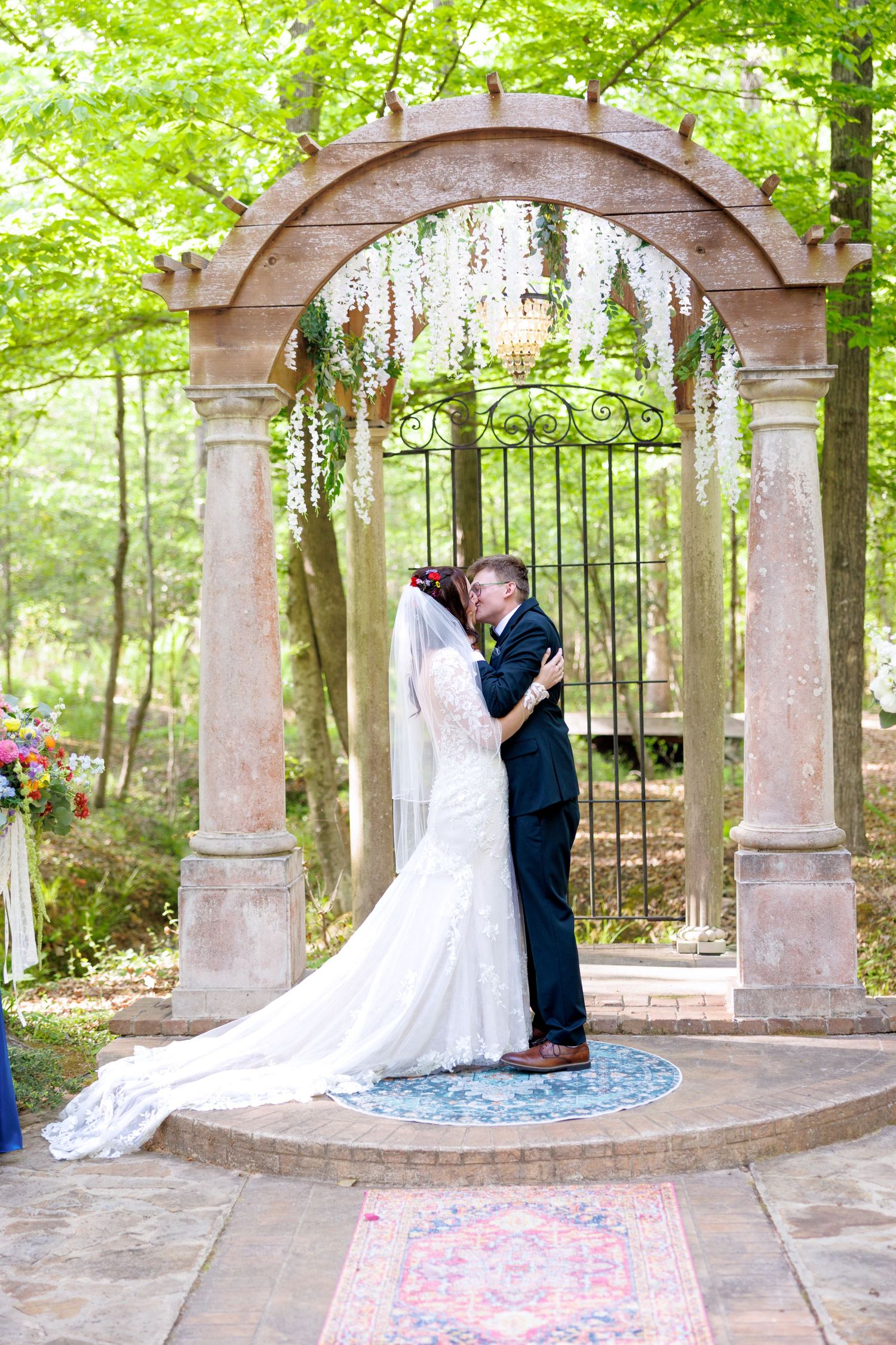 Bride and groom share first kiss under stone arch with wisteria at outdoor forest wedding ceremony.