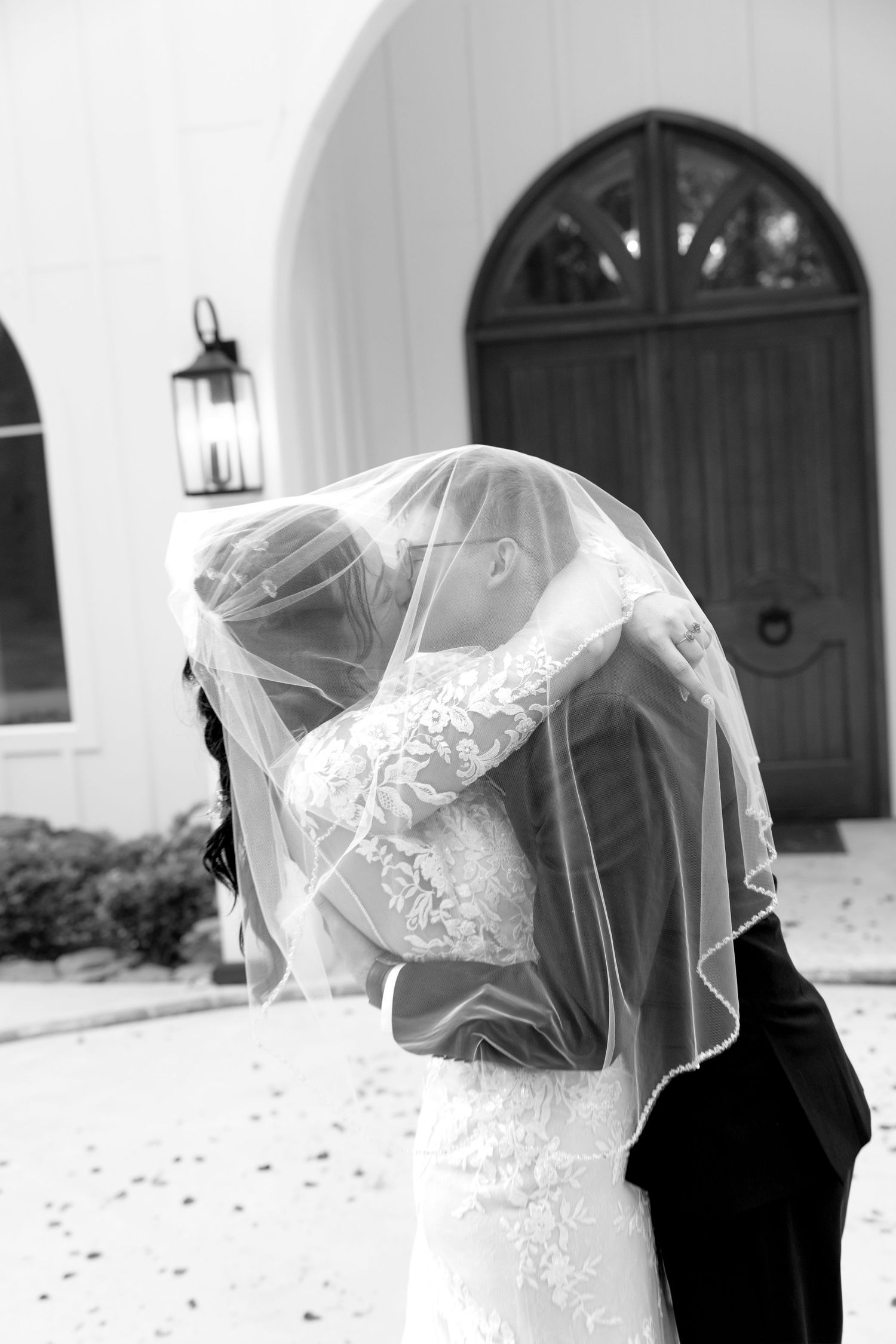 Black and white photo of bride and groom sharing a kiss under a flowing veil outside an arched church door.