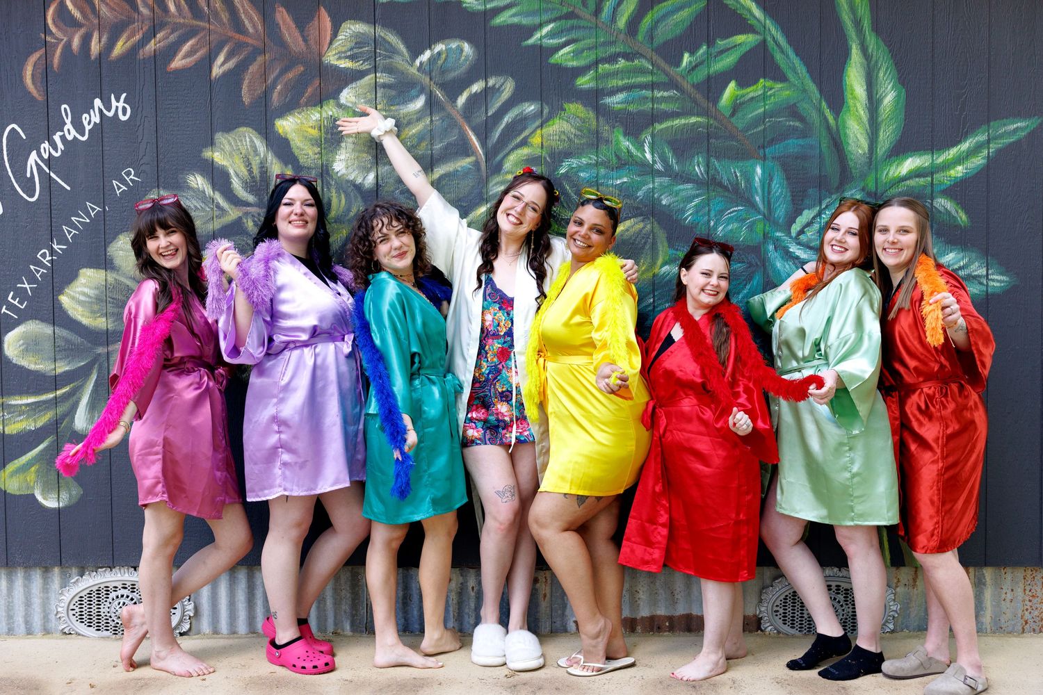Eight women in colorful satin robes pose joyfully in front of a vibrant floral mural at a bachelorette party.