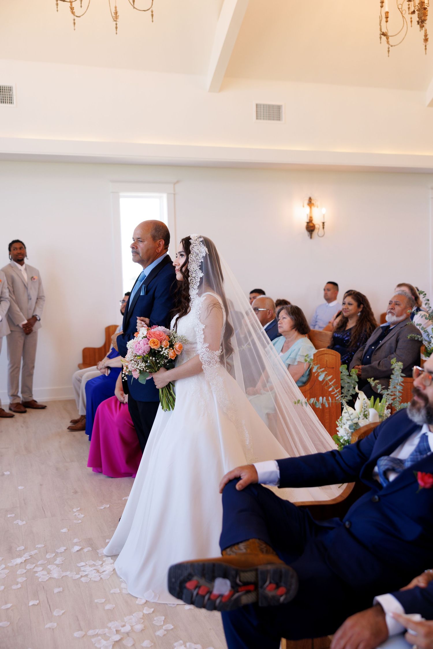 Wedding ceremony in progress with guests seated in wooden pews and rose petals scattered on the floor.