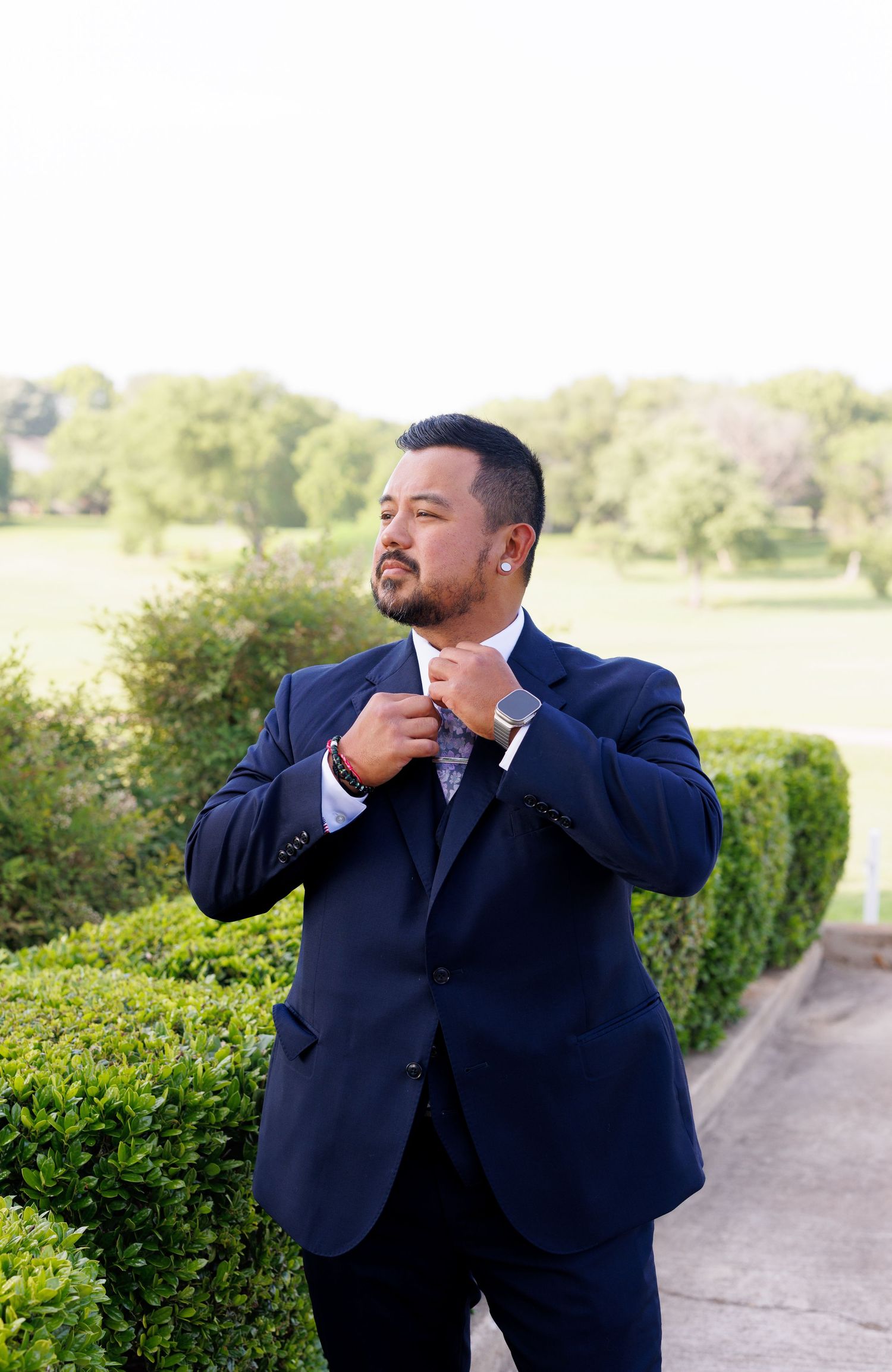 Man in navy blue suit adjusting tie outdoors at a garden venue with green hedges and trees in background.