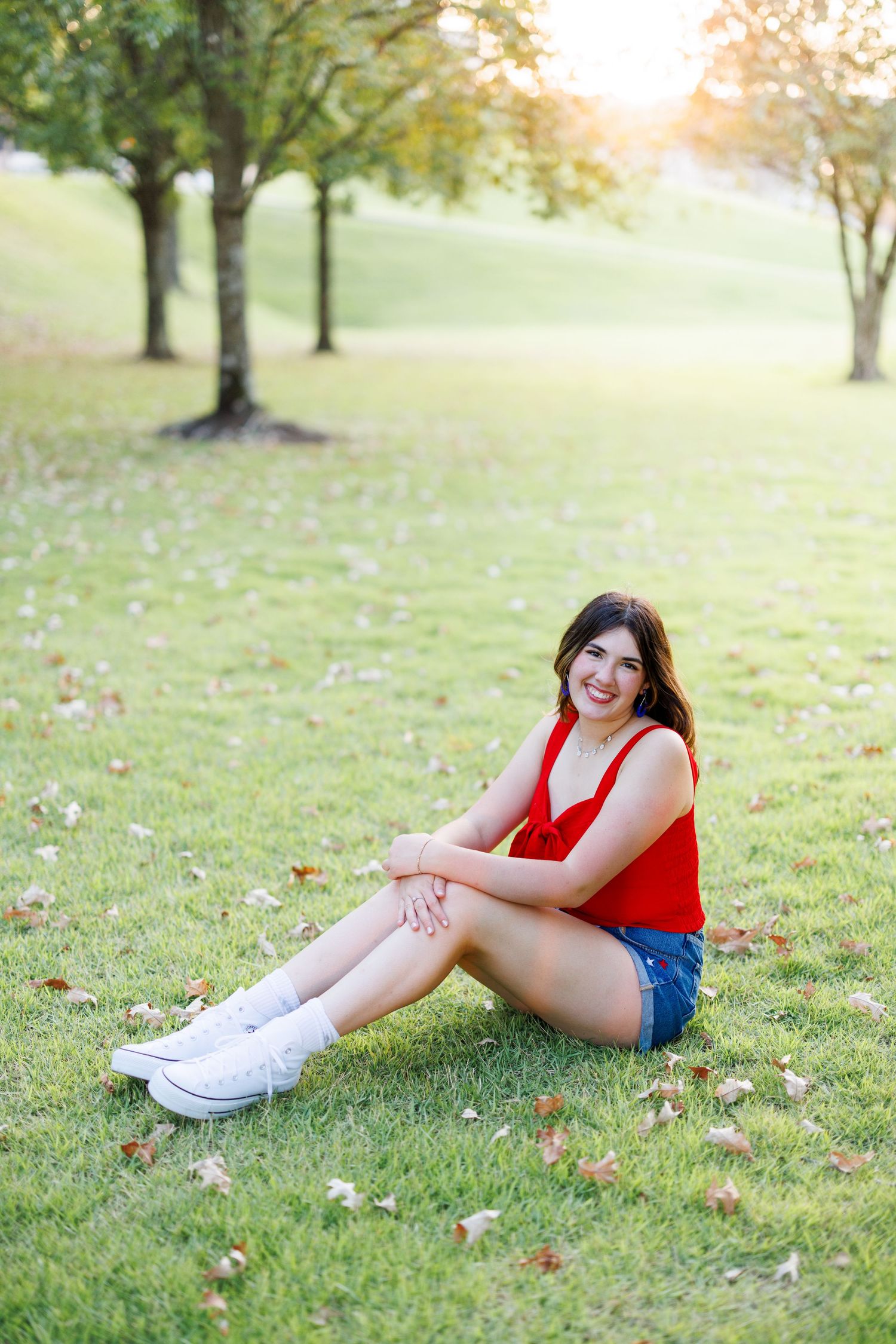 Person in red top and denim shorts relaxing on grassy lawn wearing white sneakers.
