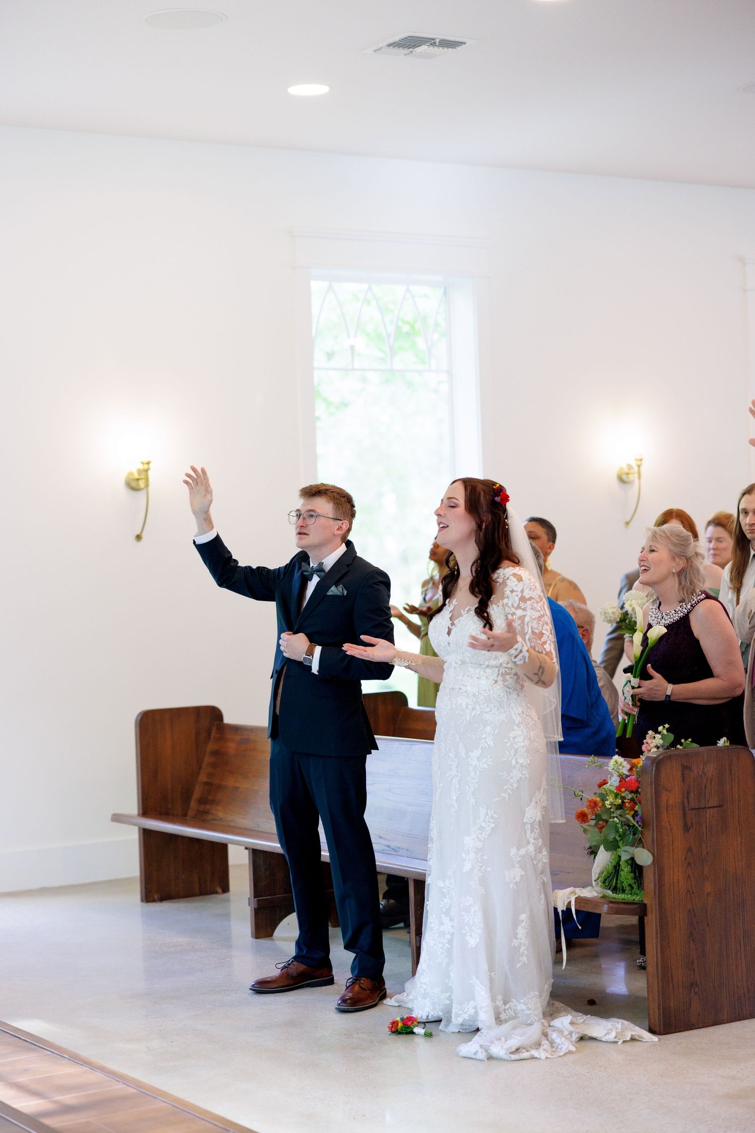 Bride and groom raise hands in worship during their wedding ceremony inside a bright white chapel with wooden pews.