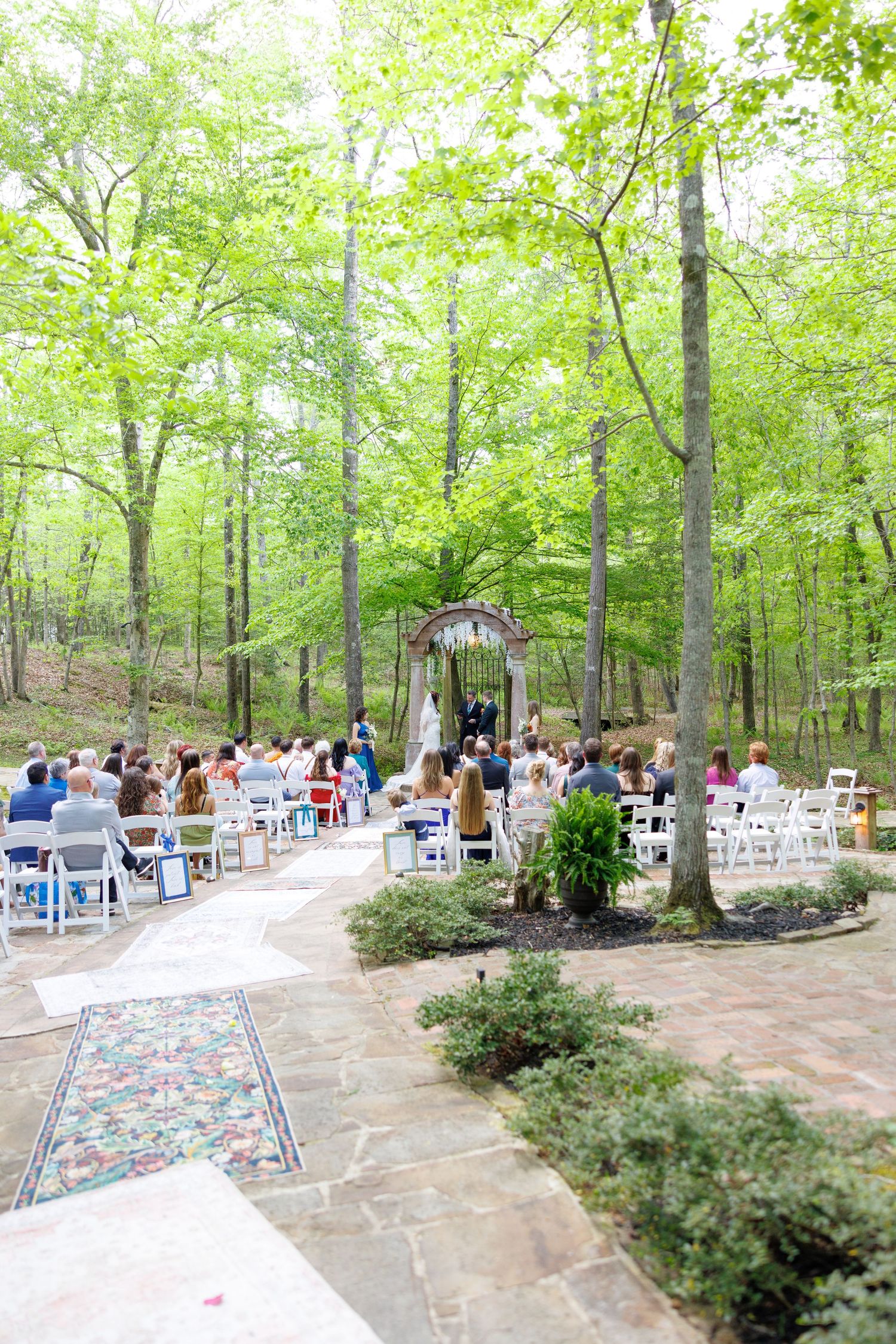An elegant outdoor wedding ceremony set among lush green trees with white chairs and a decorative arch at the altar.