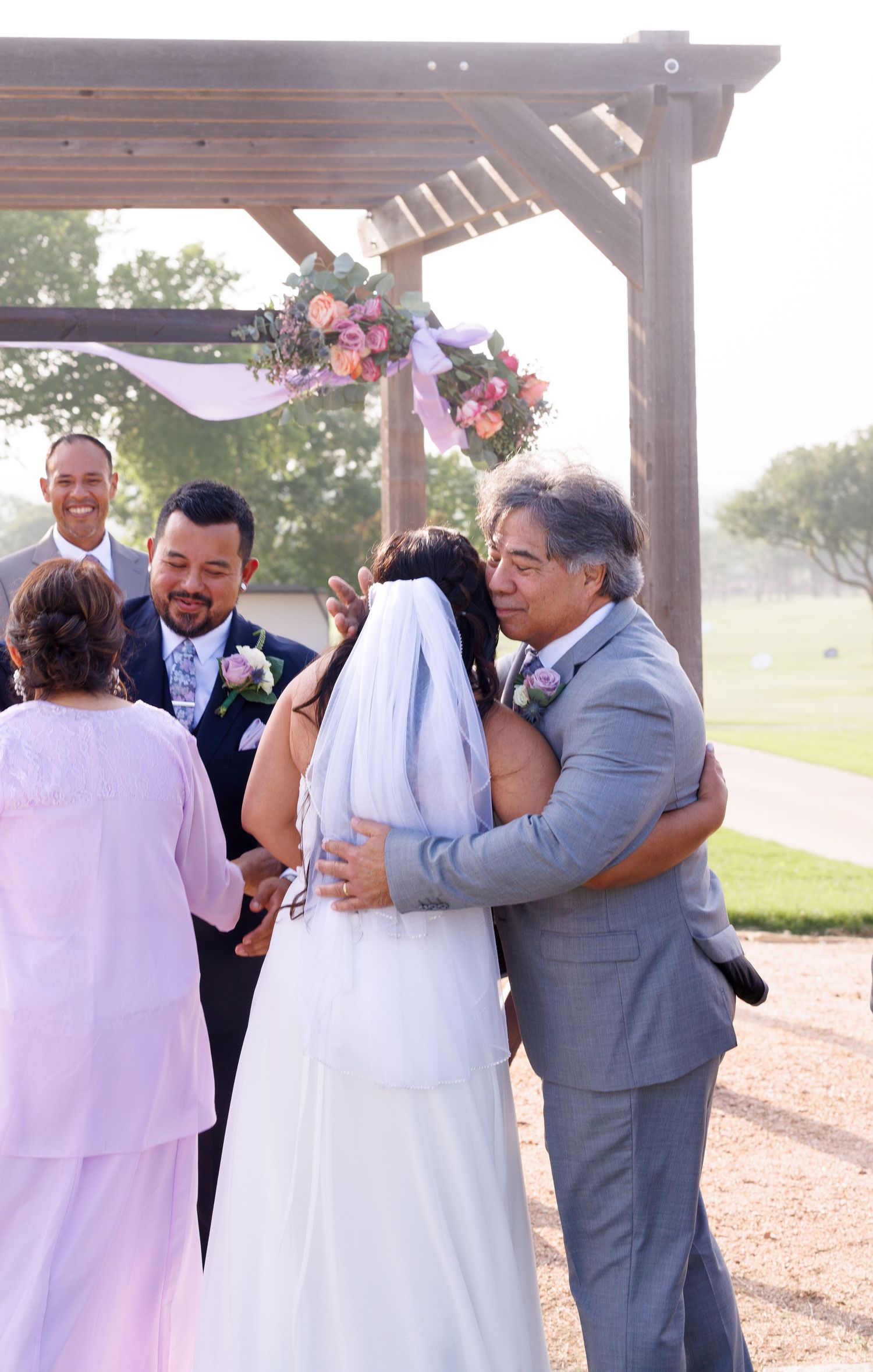 Bride and groom share a hug with family under a floral pergola during an outdoor wedding ceremony.